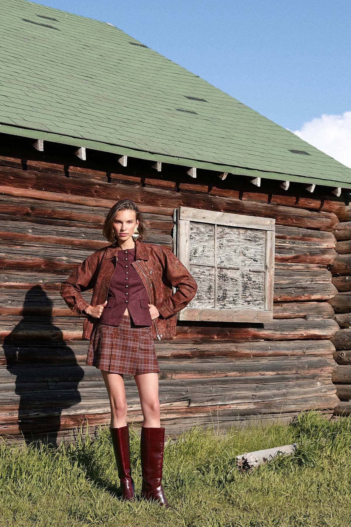 Woman wears a floral print bomber jacket, standing beside a rustic wooden cabin topped with a green roof.