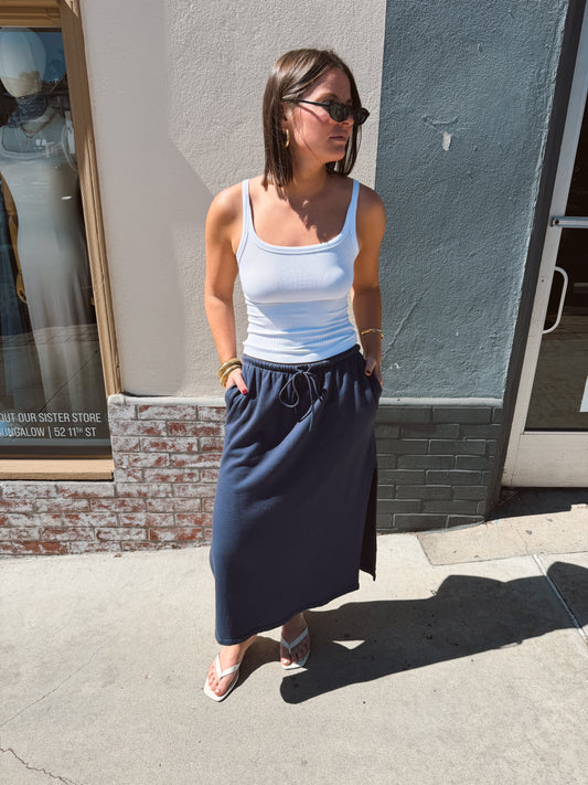 Woman in sunglasses and white tank top wears a navy pencil skirt, standing on a sunny sidewalk.