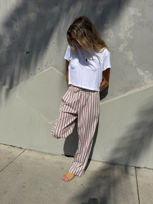 Woman wearing striped wide-leg lounge pants in stucco, hands in pockets, stands against a sunlit wall looking down.