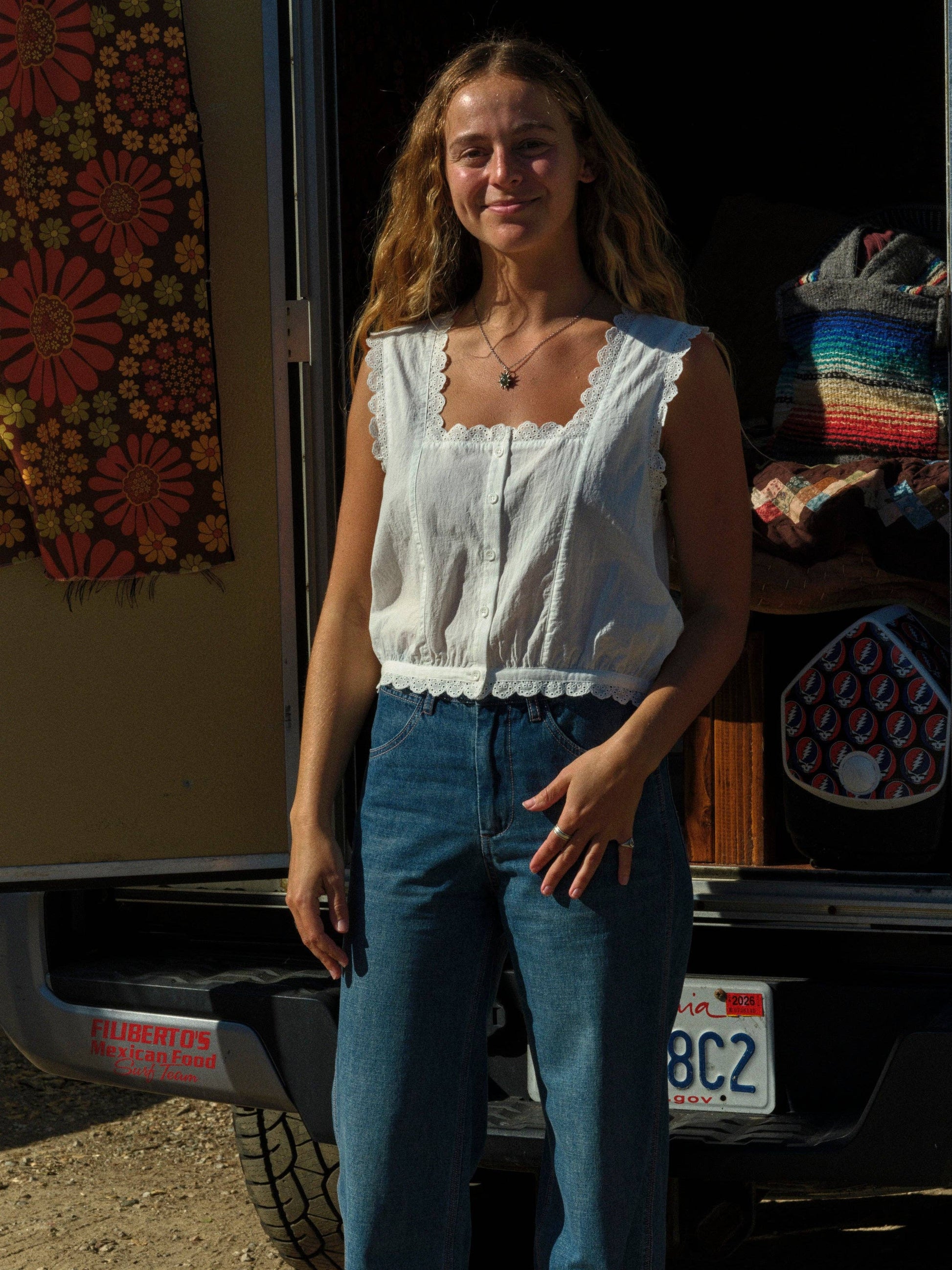Woman wearing a cotton Helena top and blue jeans stands by an open van with layered blankets and a vibrant tapestry inside.