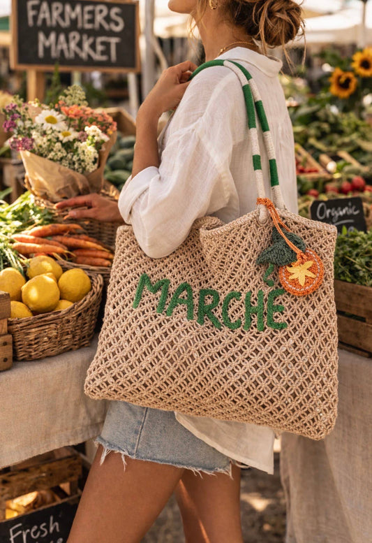 Woman carries a woven tote with raffia citrus charm at a farmers market, surrounded by fresh vegetables and flowers.