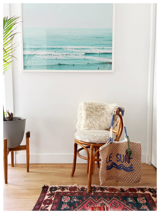 Macrame tote bag rests on a fur-cushioned chair beside a plant, beneath a framed ocean photograph.