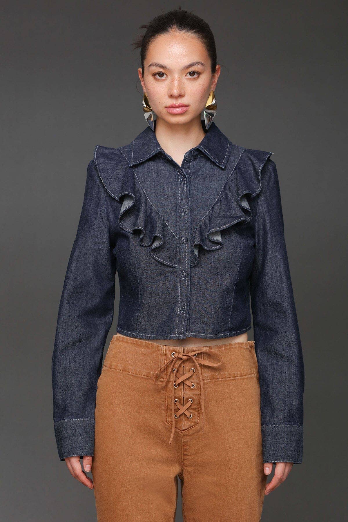 Woman wears a denim cropped shirt and tan lace-up pants, standing against a dark gray background.