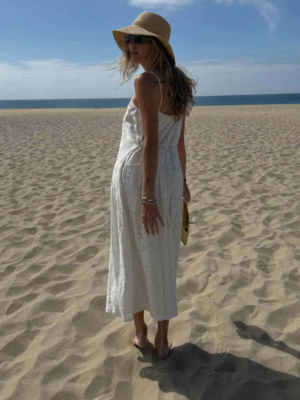 Woman wearing a white cotton midi dress and sun hat stands barefoot on sandy beach beneath a clear blue sky.