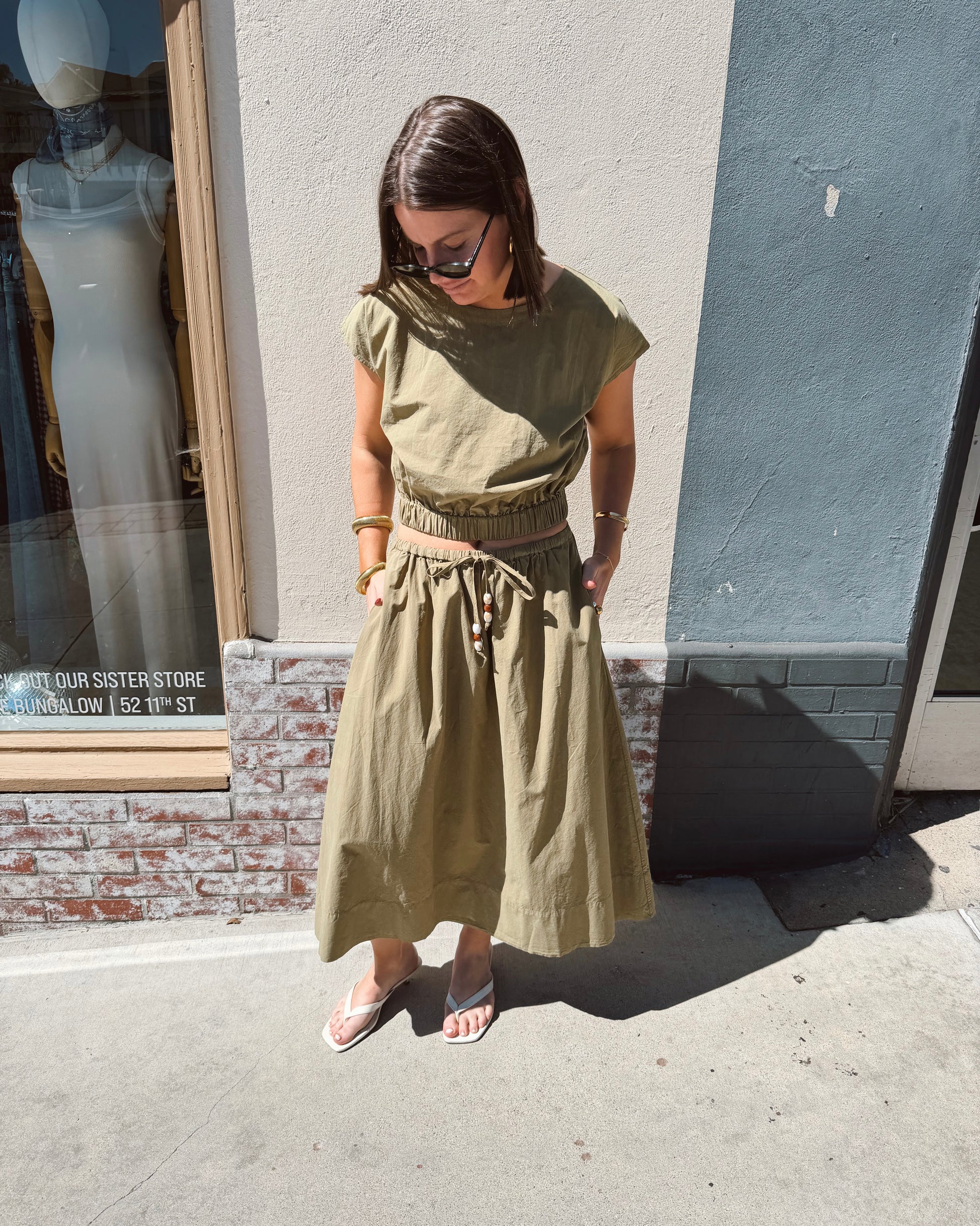 Woman wearing a dark olive tie-dye top stands on a sunny sidewalk with hands in her pockets.