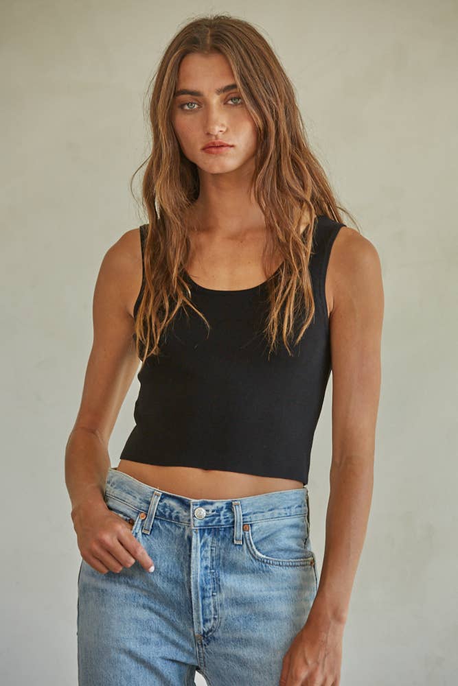 Woman with long wavy hair in a sleeveless Amanda tank top and blue denim jeans, standing before a neutral background.