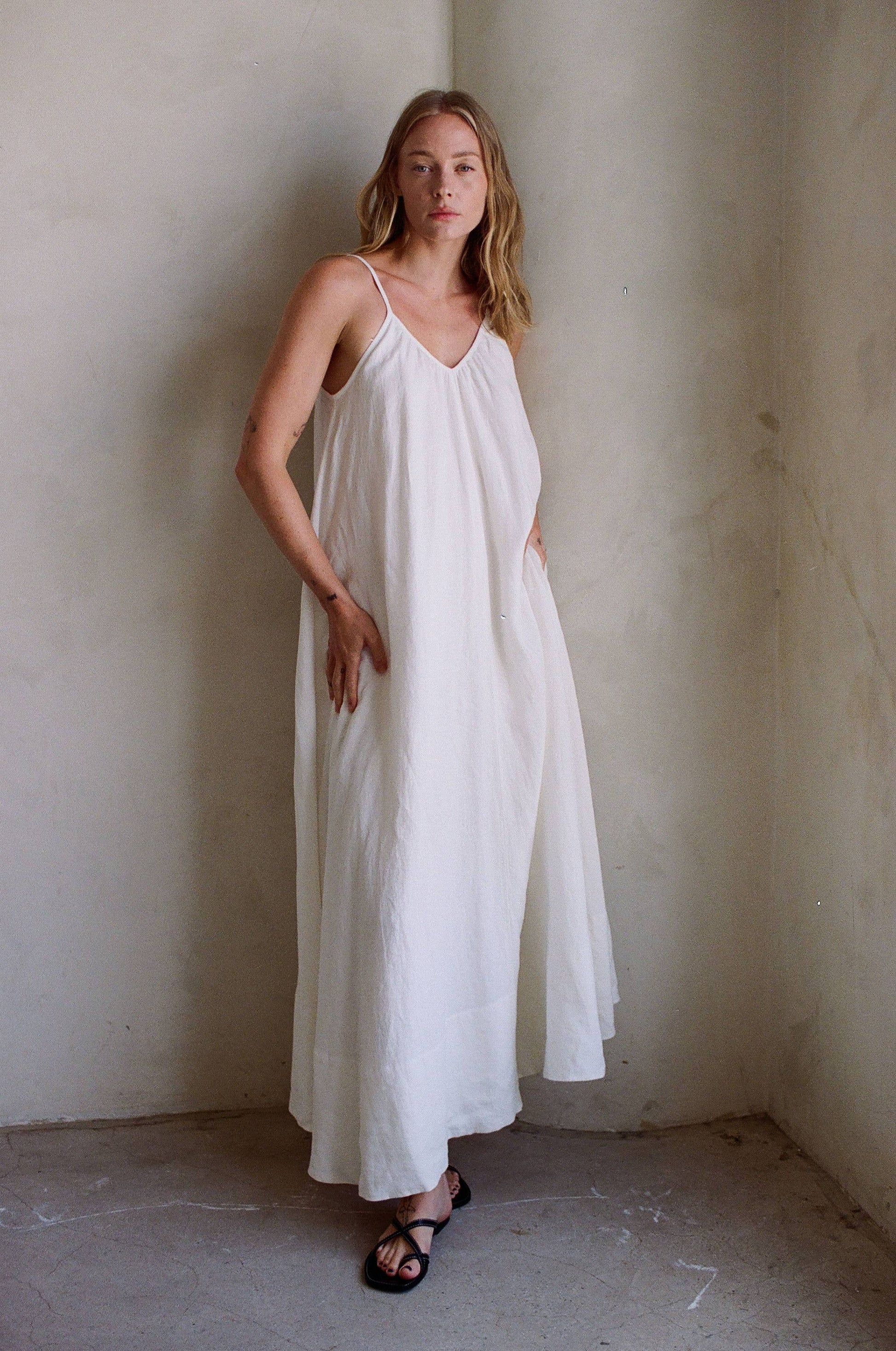 Person wearing off-white linen maxi dress with black sandals, standing by a light wall.
