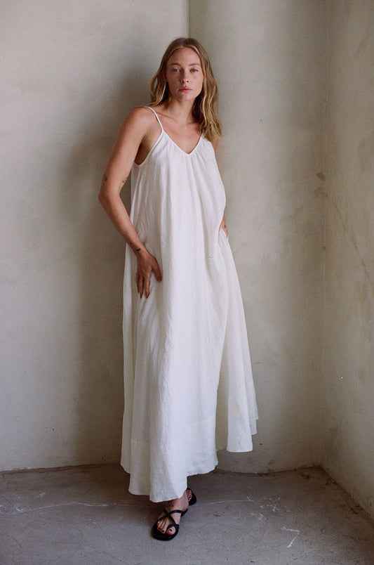 Person wearing off-white linen maxi dress with black sandals, standing by a light wall.