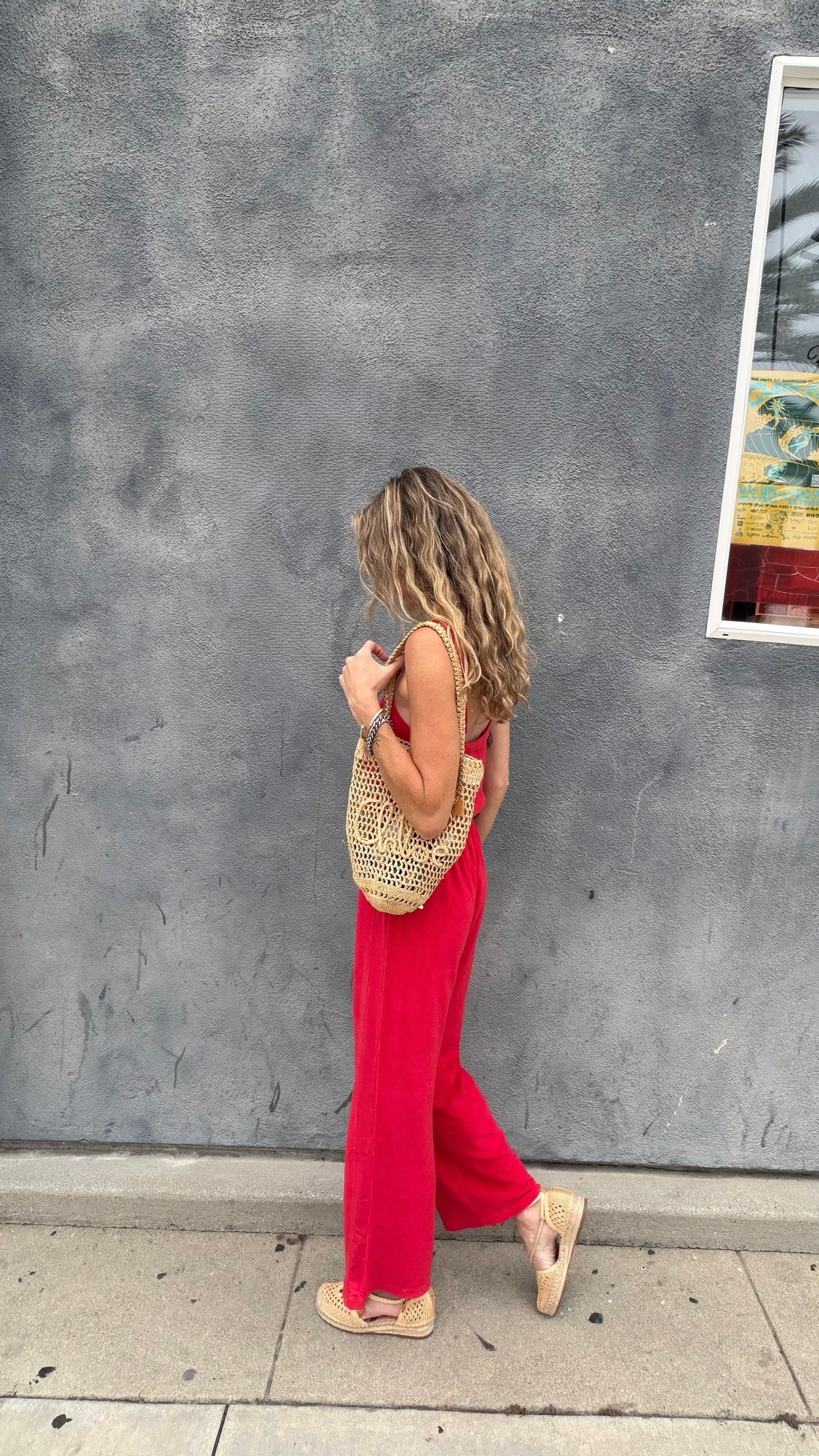 Woman in a cherry red jumpsuit walks by a gray textured wall, carrying a woven bag.