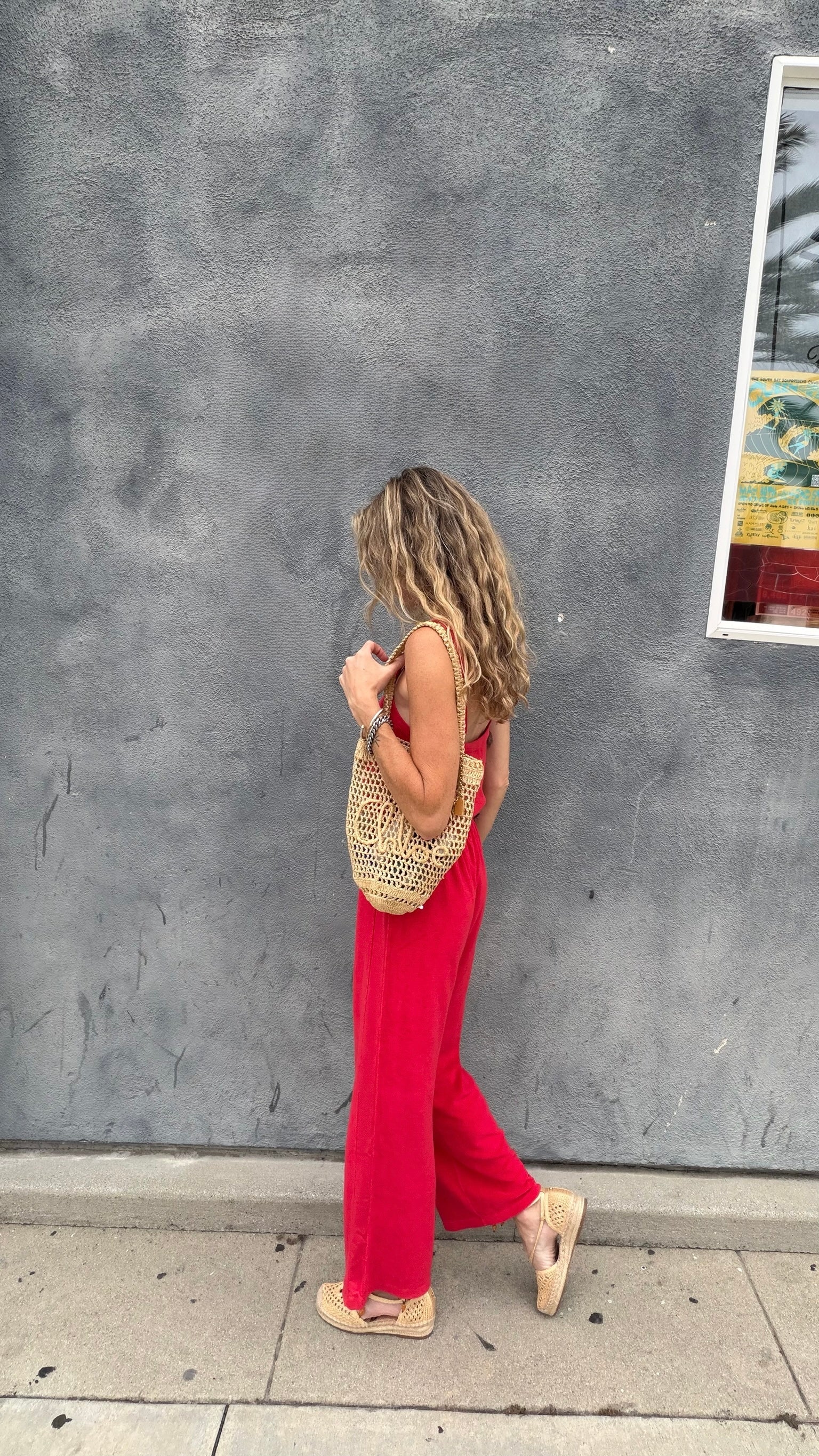 Woman in a cherry red jumpsuit walks by a gray textured wall, carrying a woven bag.