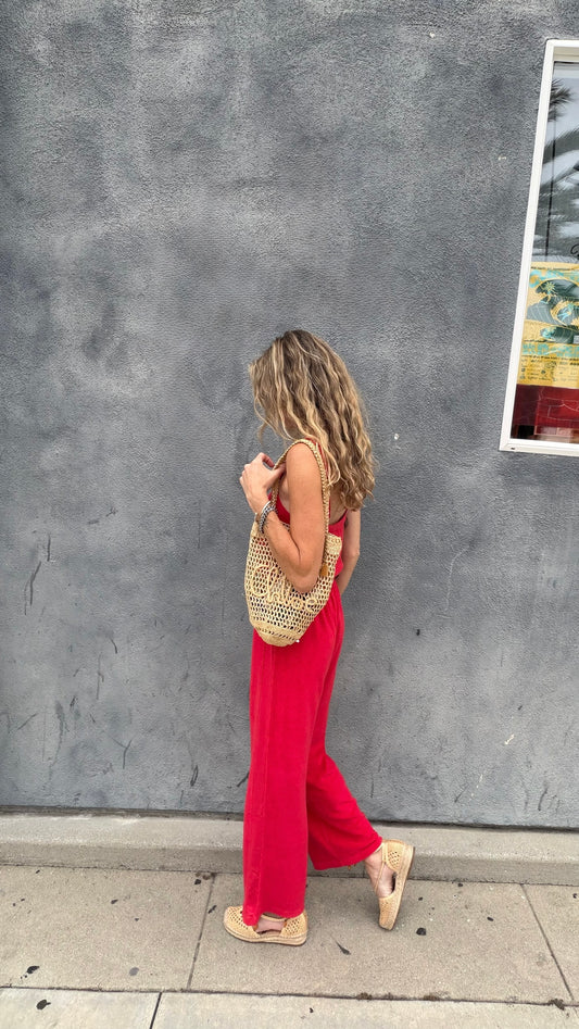 Woman in a cherry red jumpsuit walks by a gray textured wall, carrying a woven bag.