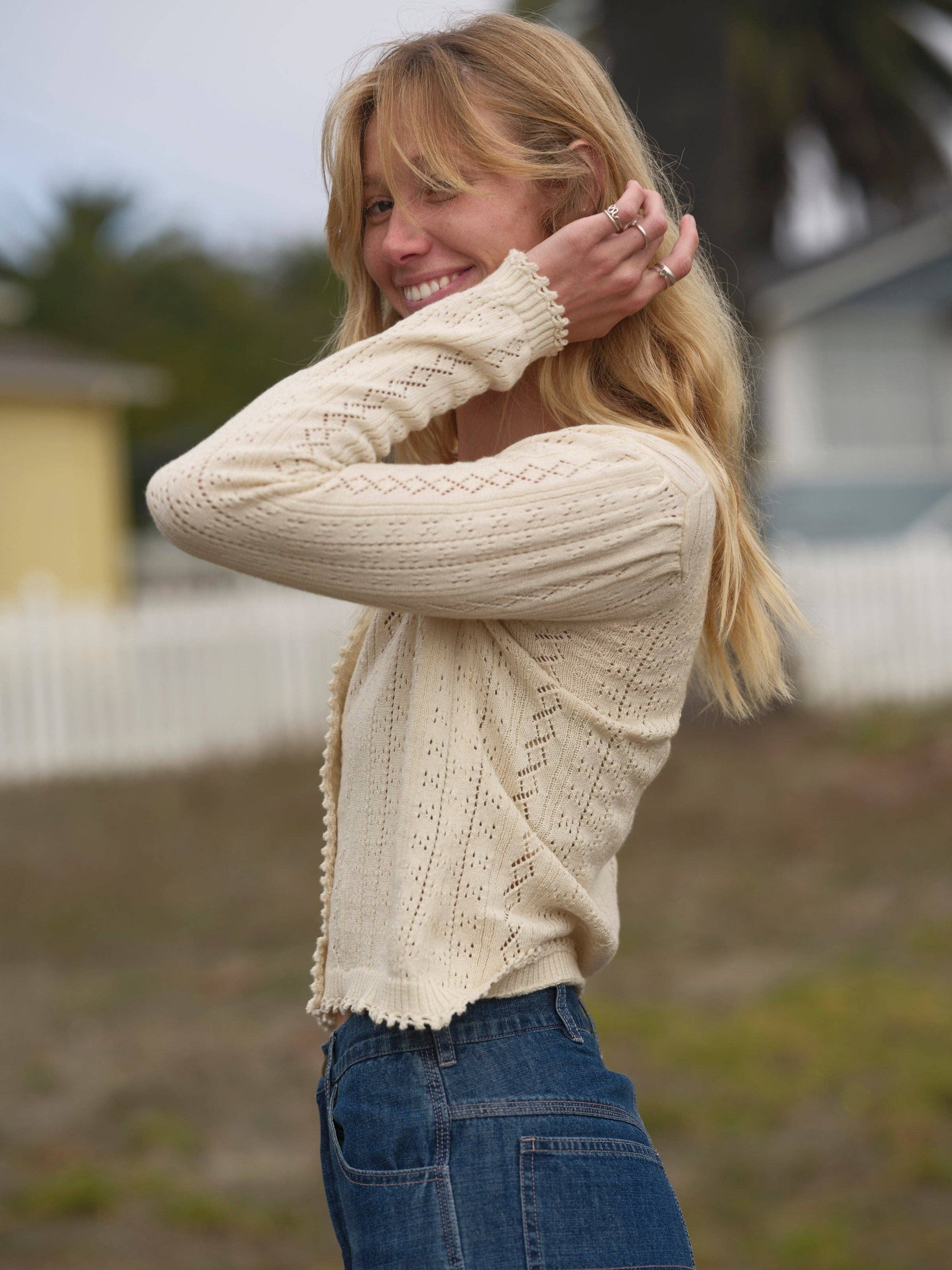 Blonde woman in a cream-colored knit cardigan stands by a white picket fence outdoors.