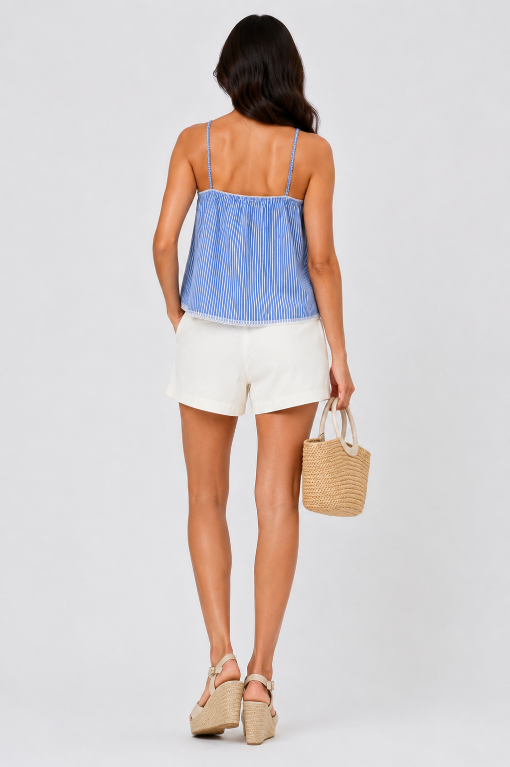 Woman in blue striped top, white shorts, wedges, and woven bag, facing away.