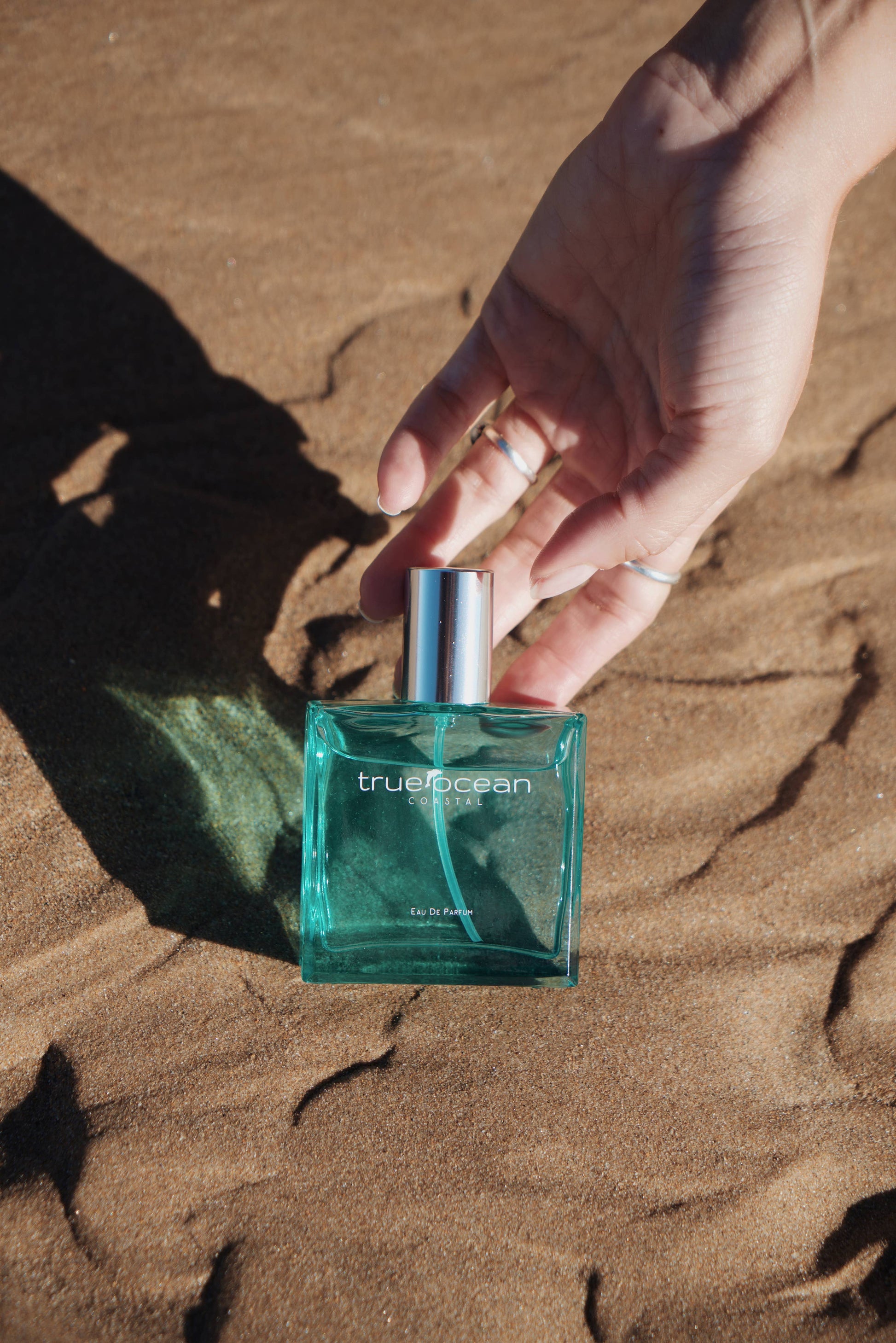 Hand reaches for turquoise glass perfume bottle on rippled sand, sunlight highlighting its ocean-inspired design.