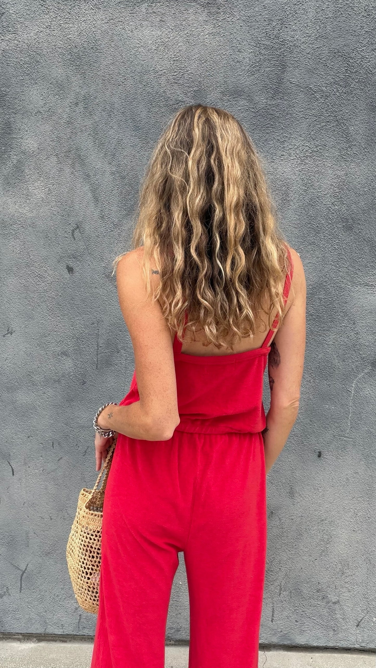 Woman with wavy blonde hair wears a cherry red jumpsuit and holds a woven tote by a gray wall.