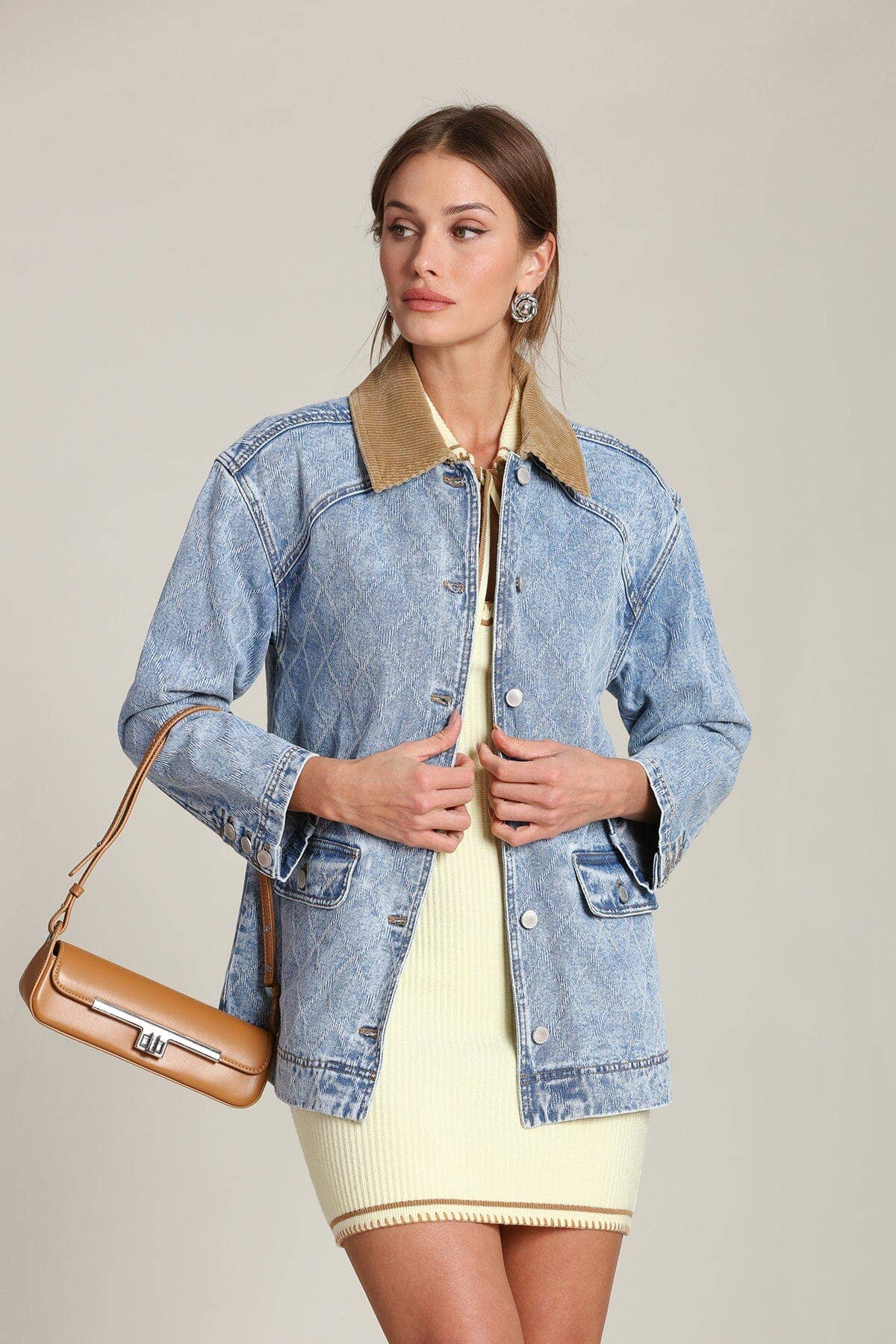 Woman wears denim barn jacket over cream dress, holding a tan handbag; neutral backdrop. Denim, cotton, and leather materials.