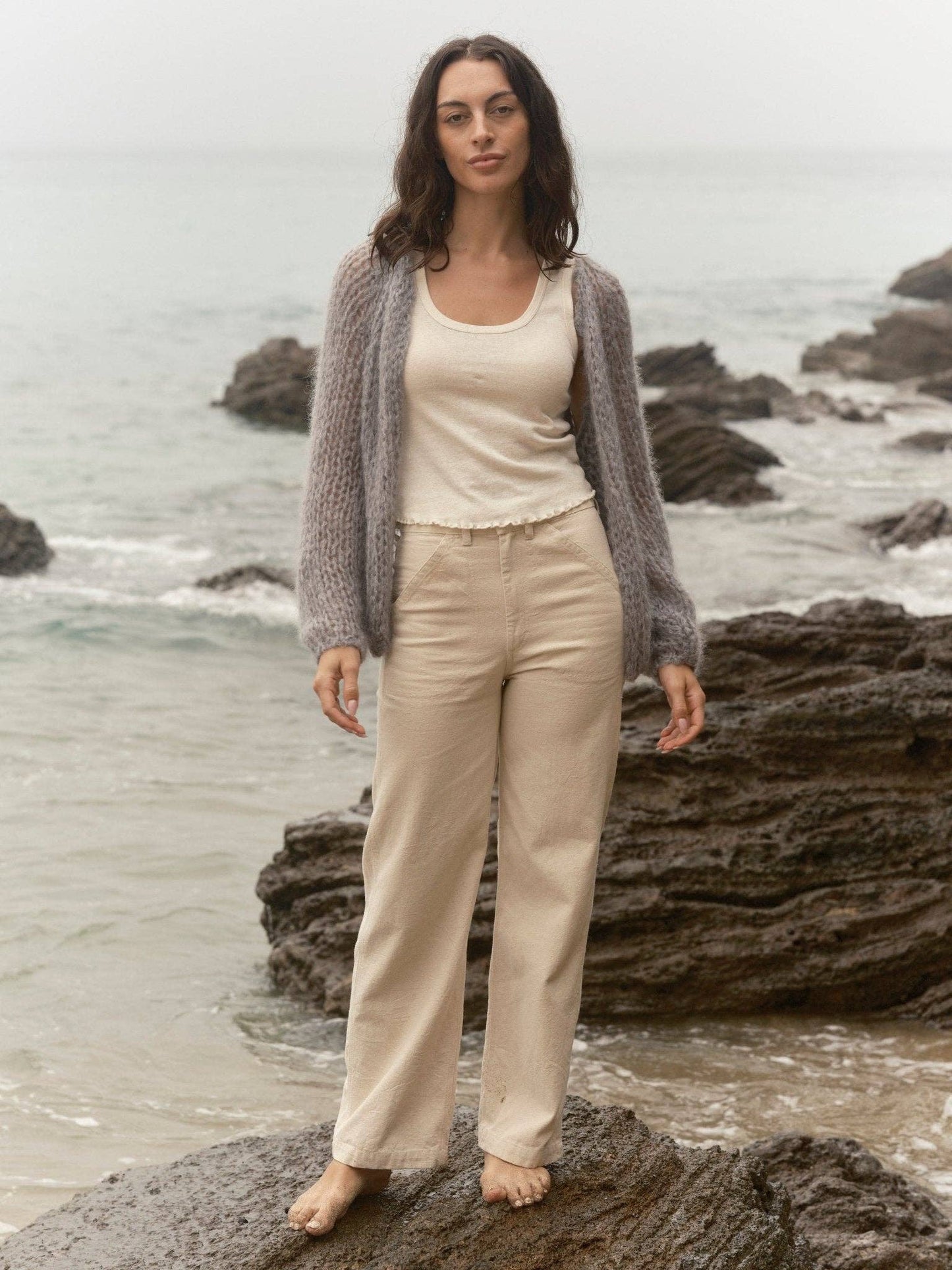 Woman in gray knit cardigan stands barefoot on seaside rocks, waves and cloudy sky in the background.