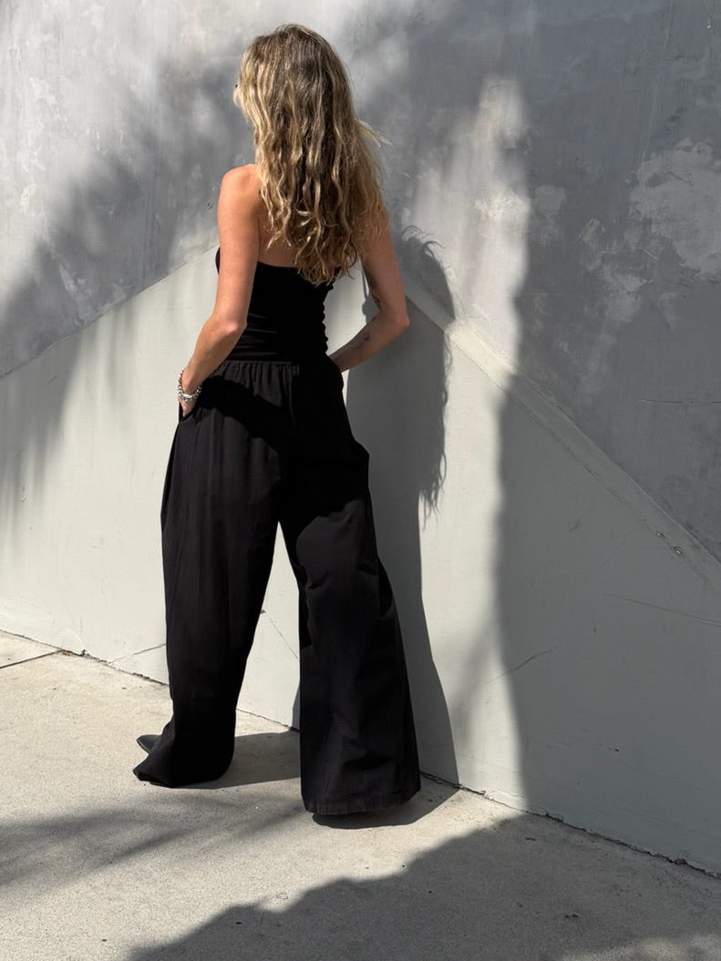 Woman in a black jumpsuit, fabric draping softly, with sunlight creating patterns on a gray wall in the background.