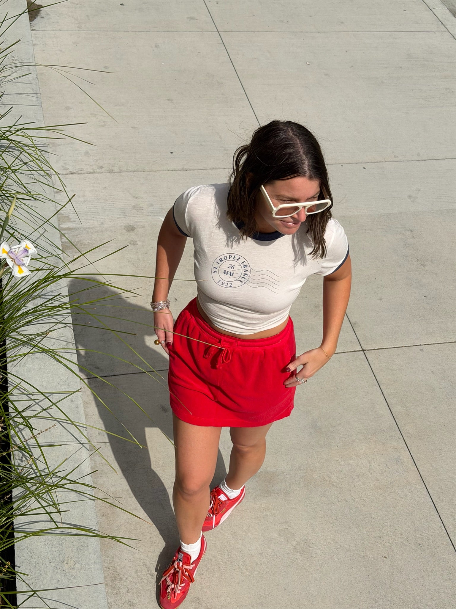 Woman in a white crop top, vermillion terry mini skirt, and white sunglasses walking on a sunlit sidewalk.