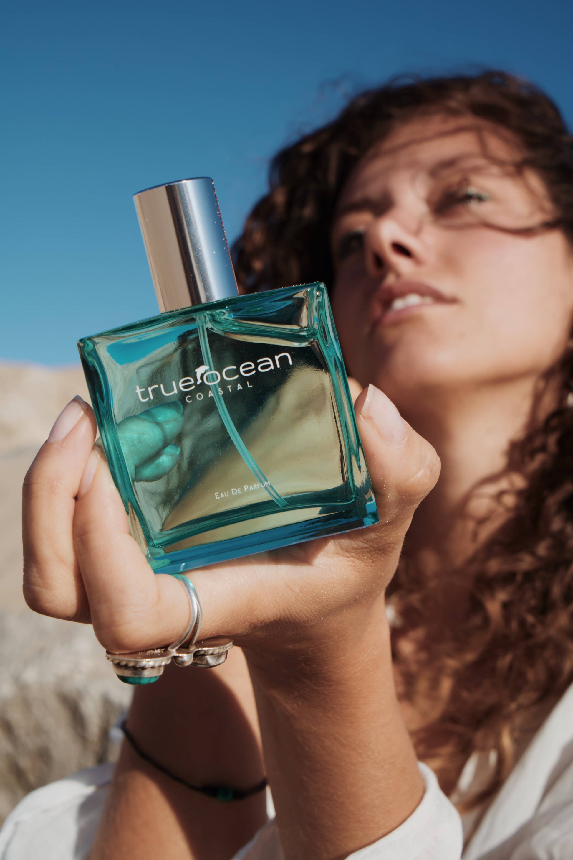 Woman holding a clear glass Coastal perfume bottle outdoors under a blue sky, suggesting a fresh, beach-inspired use.