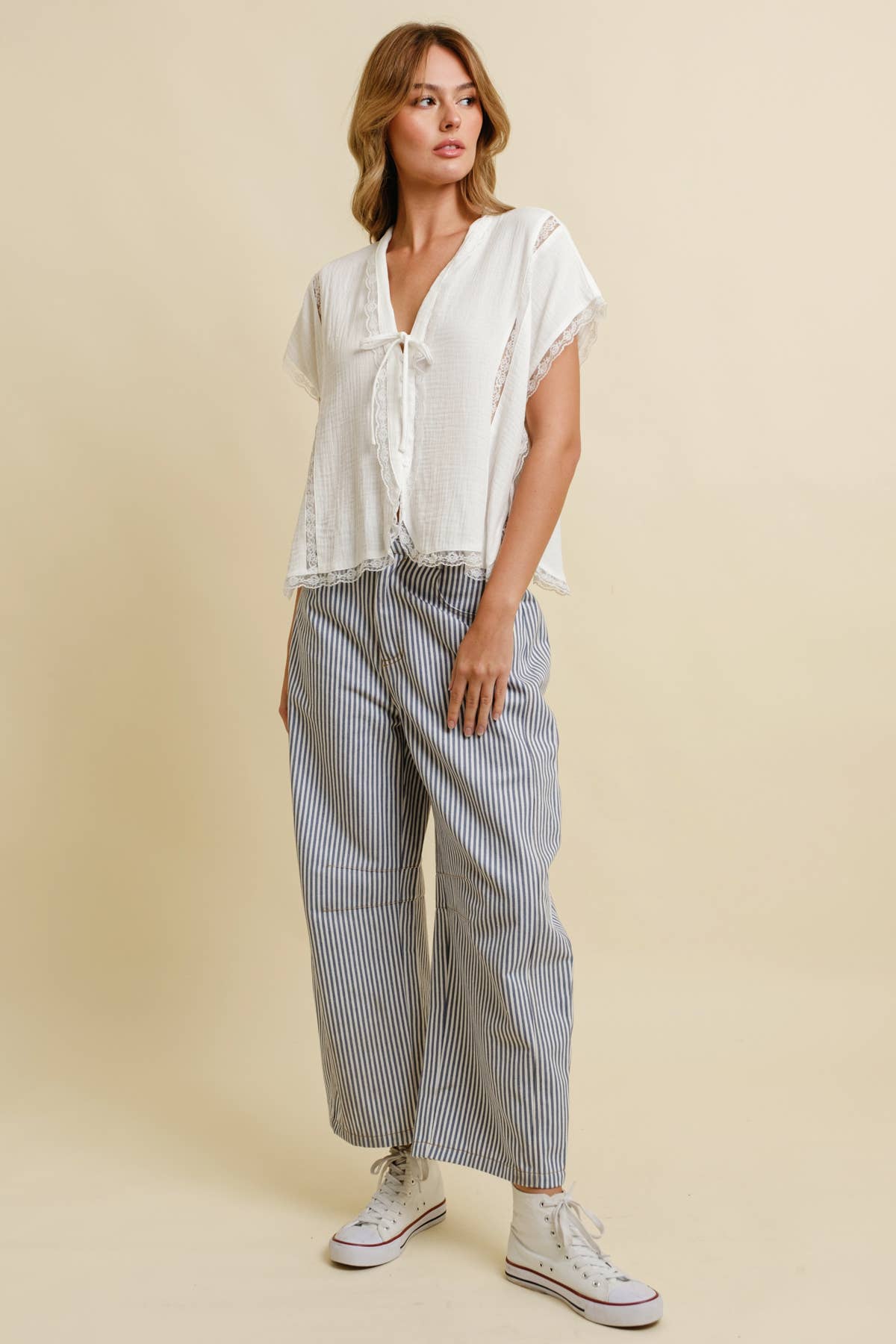 Woman wears a light top, striped wide-leg pants, and white sneakers, standing against a beige background.
