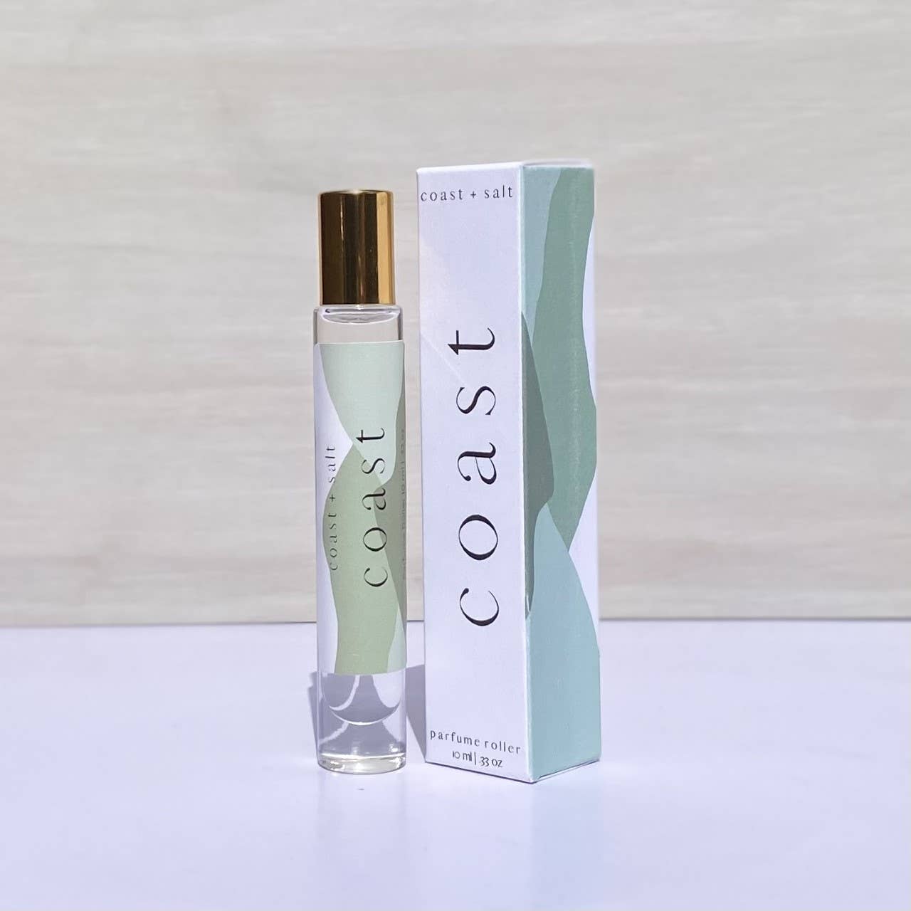 Sandalwood Coast+Salt perfume roller displayed next to its box featuring an abstract green and white design.