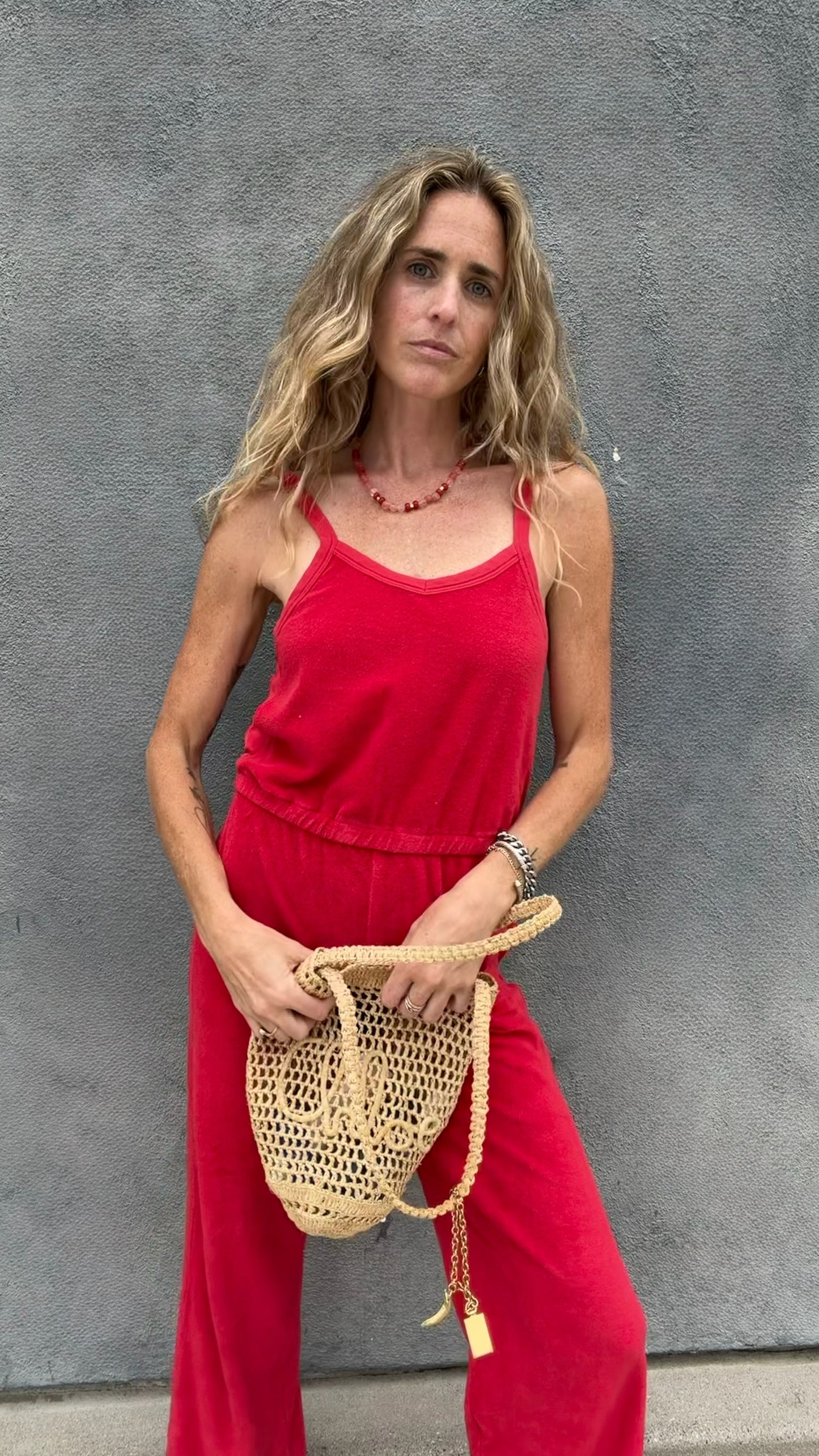 Woman wears a cherry red jumpsuit and holds a woven bag, standing in front of a gray textured wall.