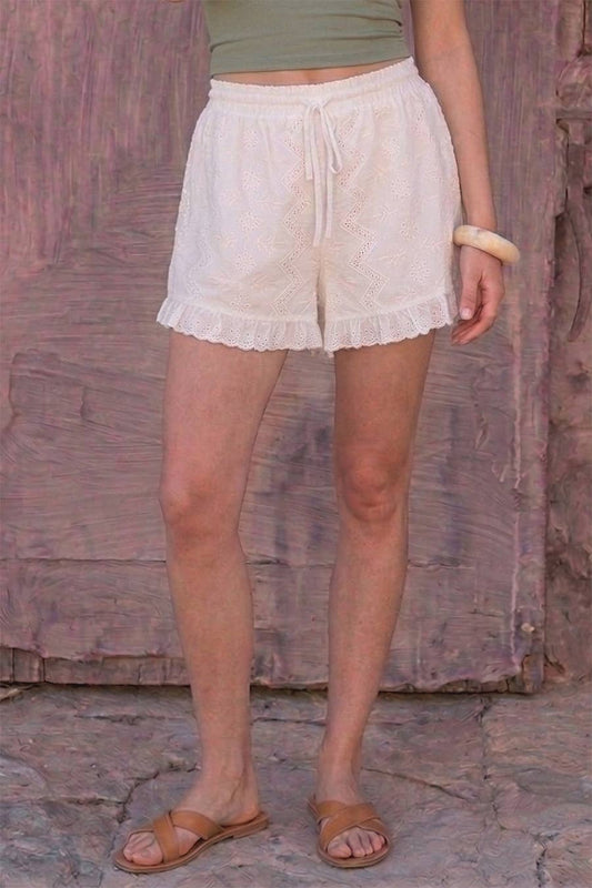 Woman in off-white ruffle shorts, green top, tan sandals, and bangle standing by a textured wall.