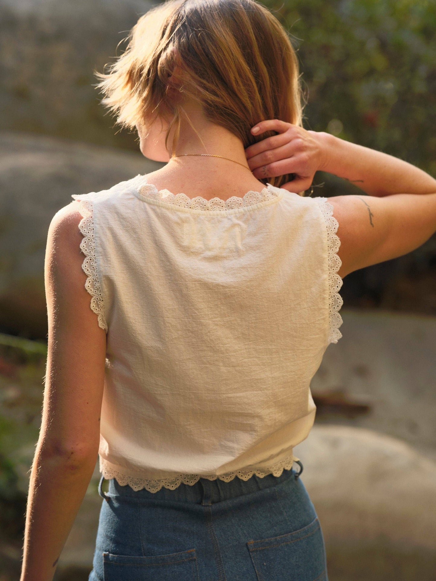 Person outdoors in a cream cotton top and blue denim jeans, seen from behind, hand touching hair.