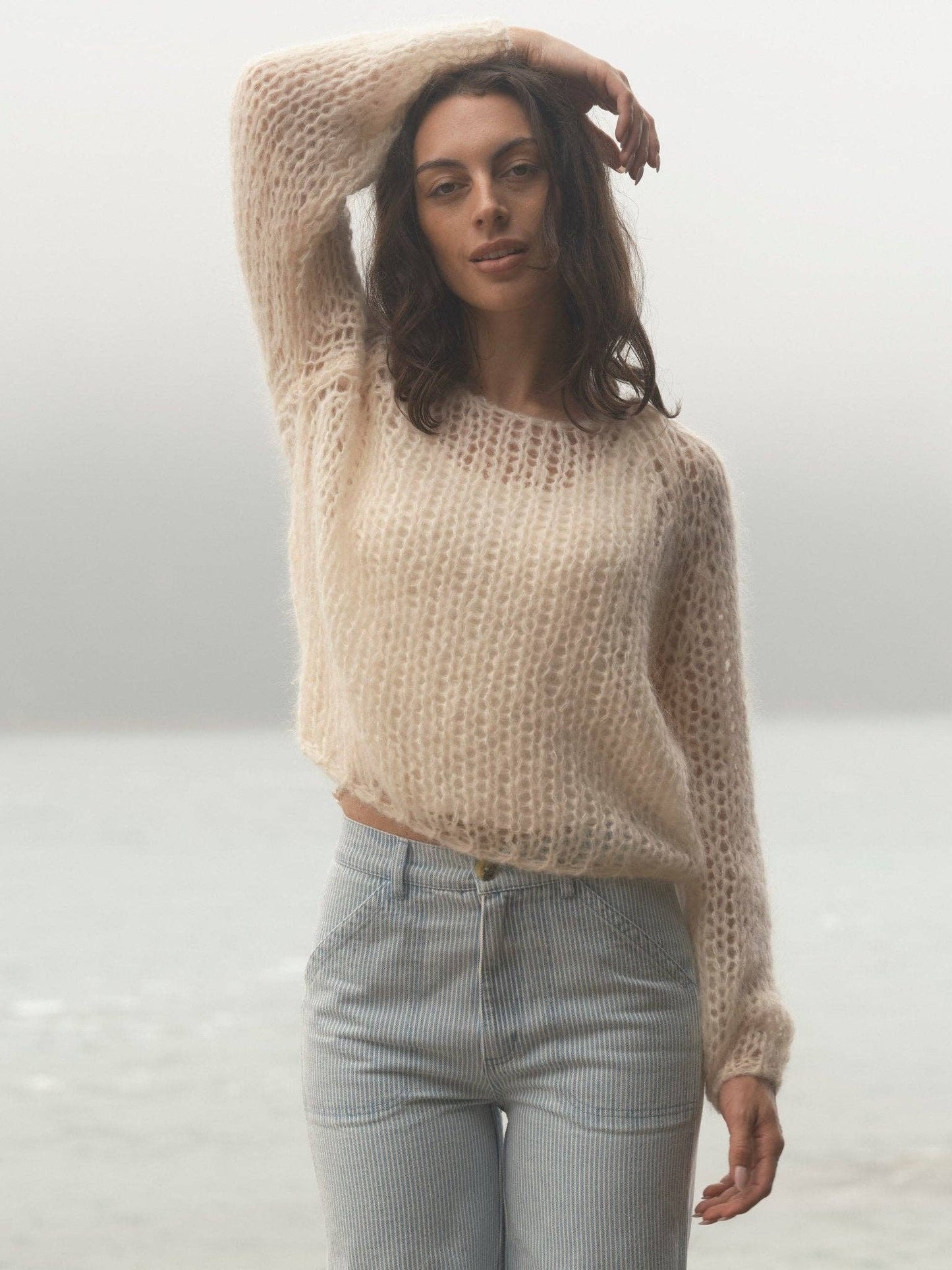 Woman by misty water wears a loose cream sweater and light jeans, arm lifted overhead. Sweater is soft knit.