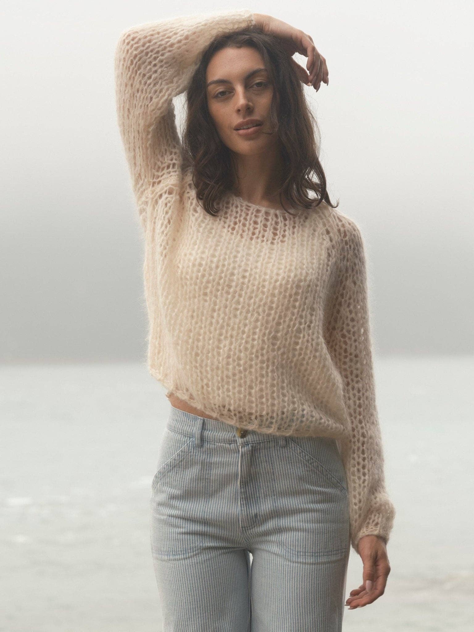 Woman by misty water wears a loose cream sweater and light jeans, arm lifted overhead. Sweater is soft knit.