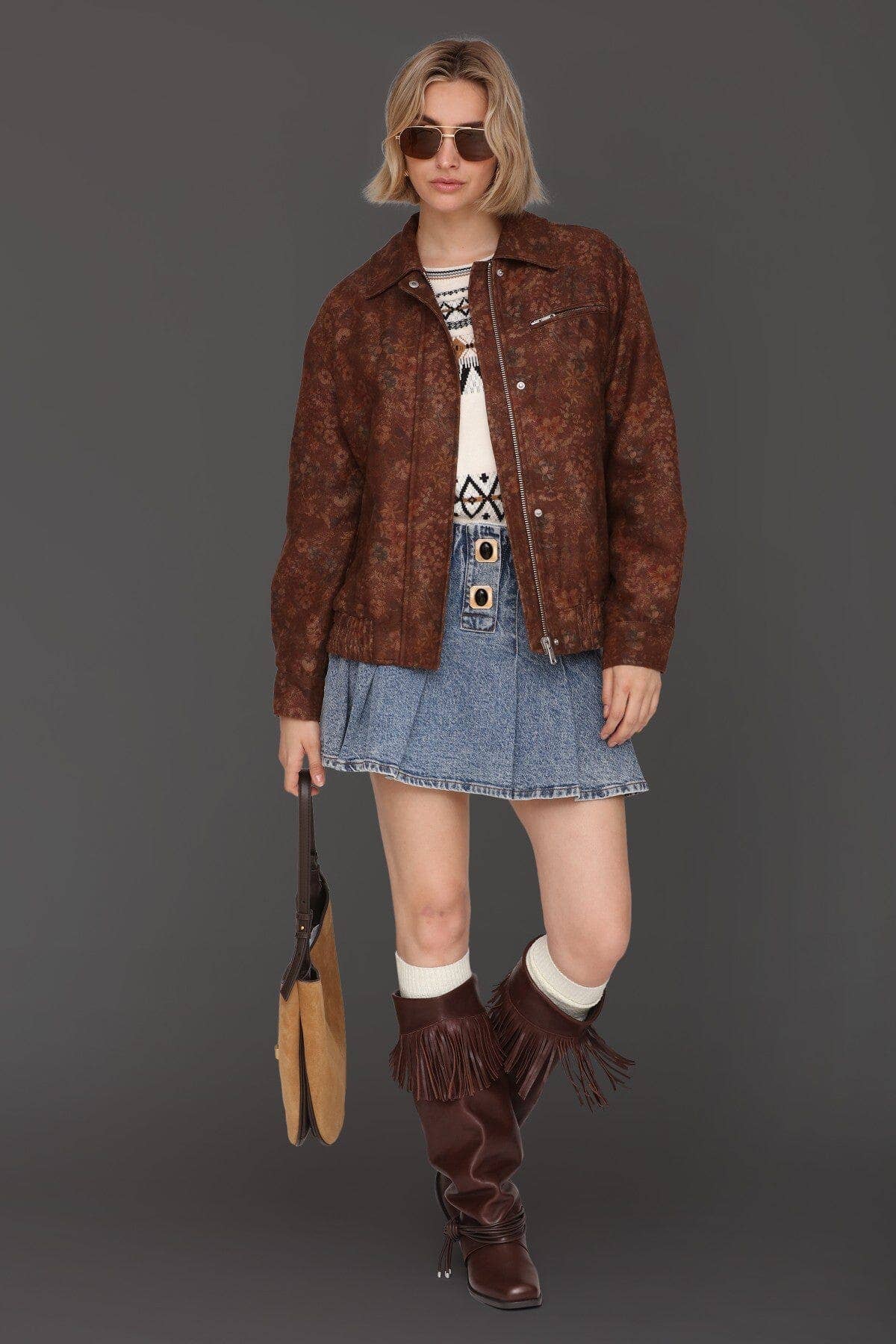 Person in sunglasses, floral bomber jacket, patterned sweater, blue denim skirt, black boots, and carrying a handbag.