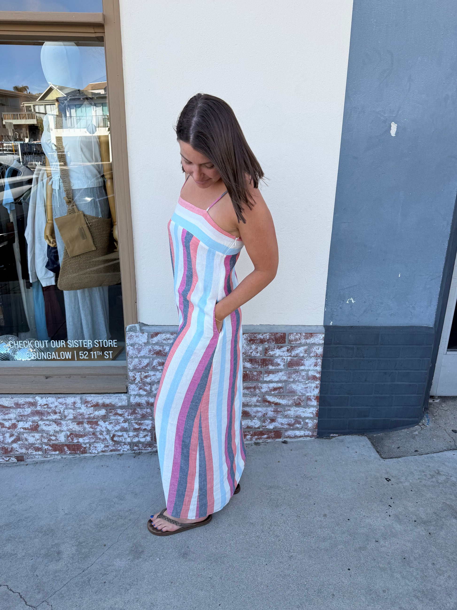 Woman wearing a striped multi-color maxi dress stands on the sidewalk with hands in her pockets.