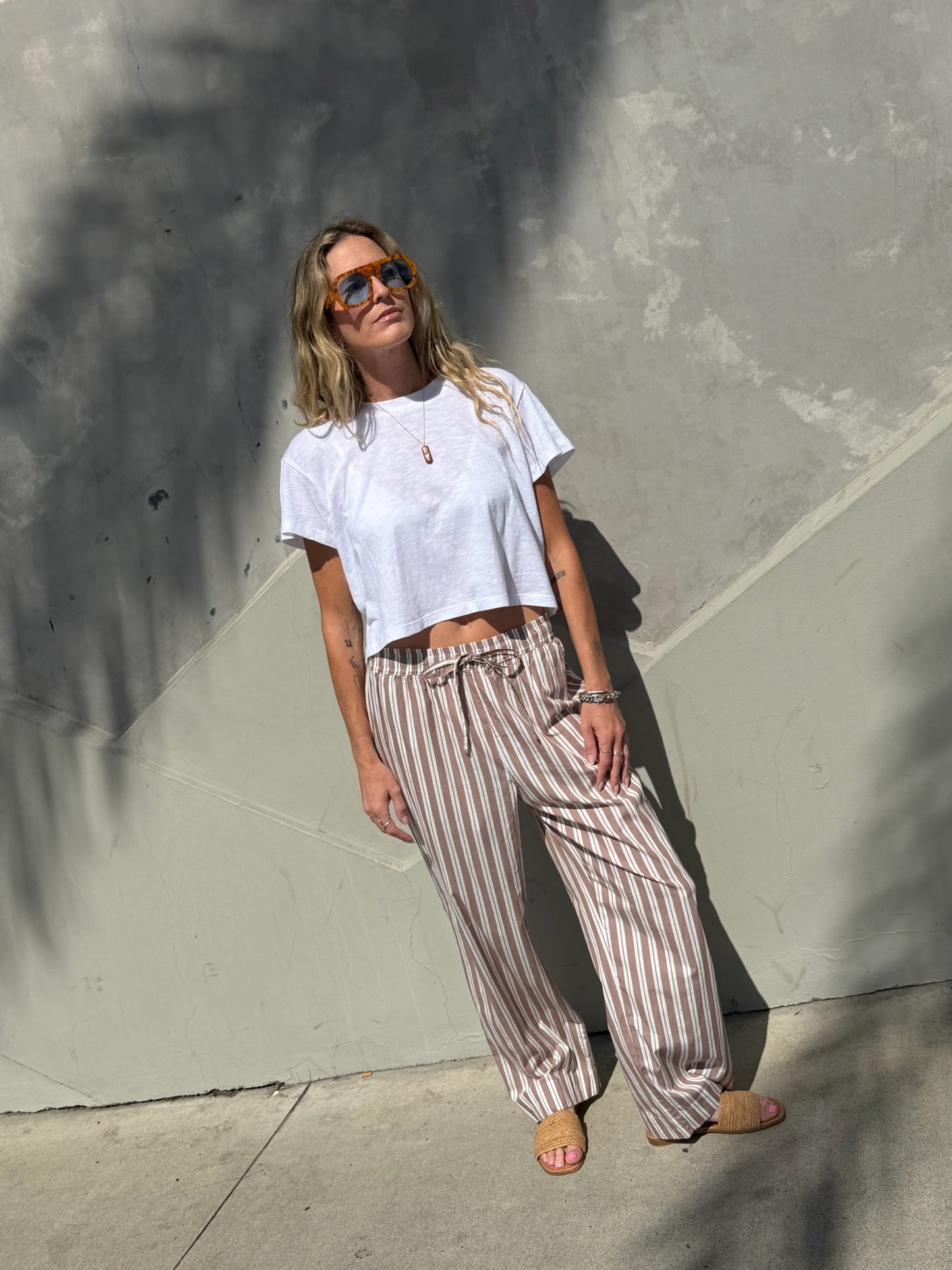 Woman wearing striped wide-leg lounge pants in stucco and sunglasses stands by a sunlit textured concrete wall.