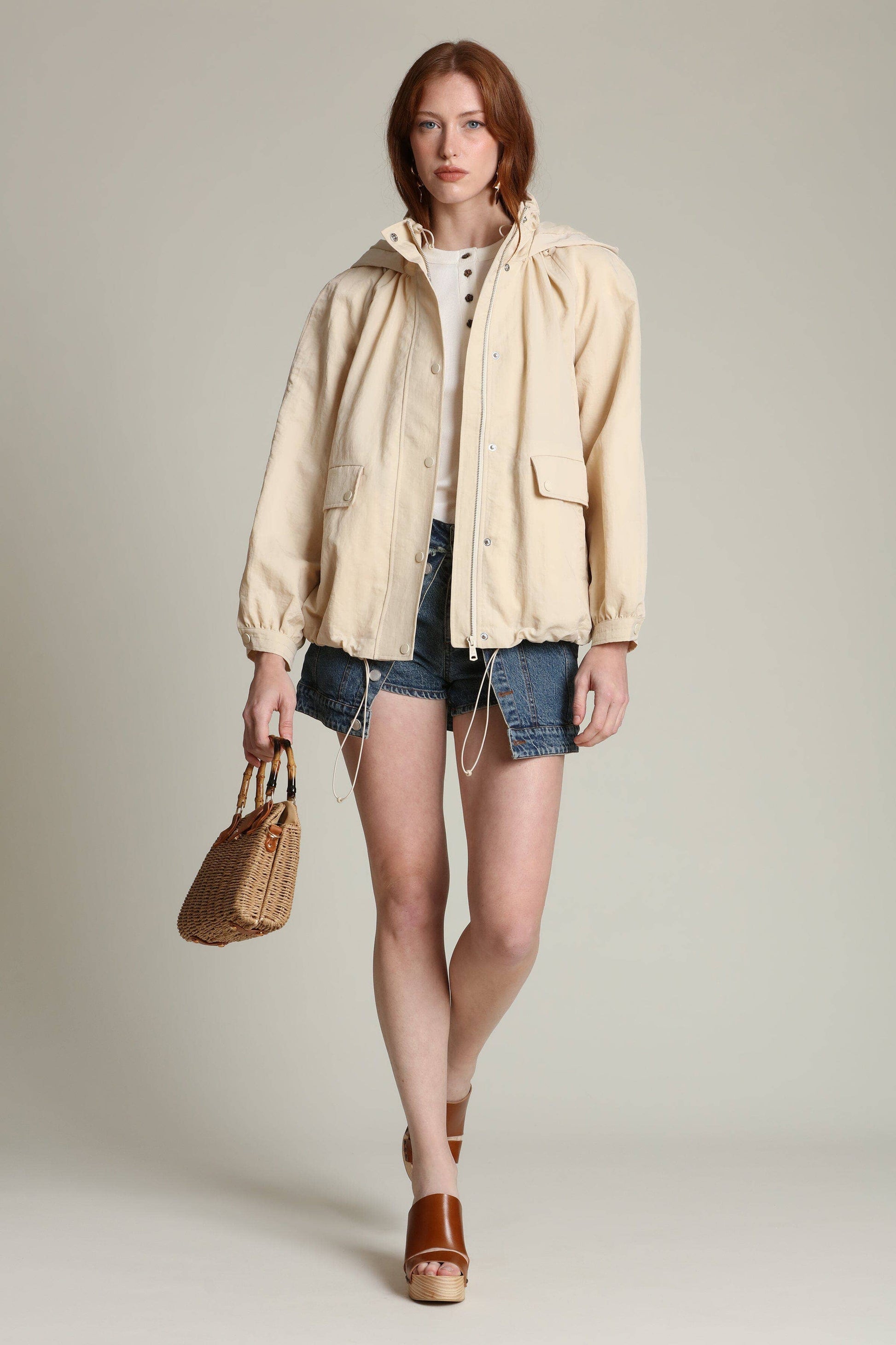Woman wearing a balloon sleeve windbreaker, white shirt, denim shorts, sandals, and carrying a woven handbag.