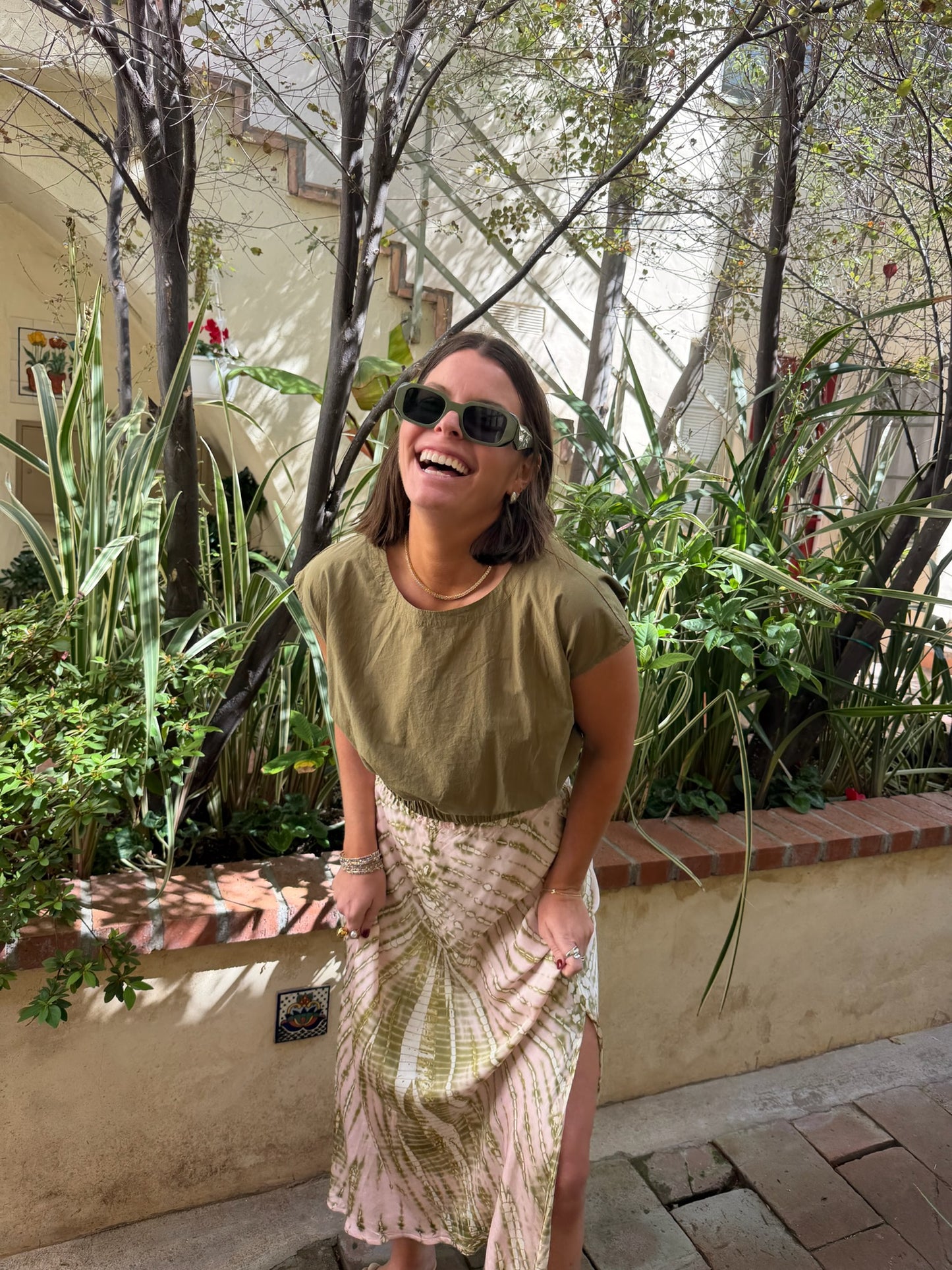 Woman in sunglasses laughs outdoors, wearing a dark olive top made of soft fabric, suitable for casual use.