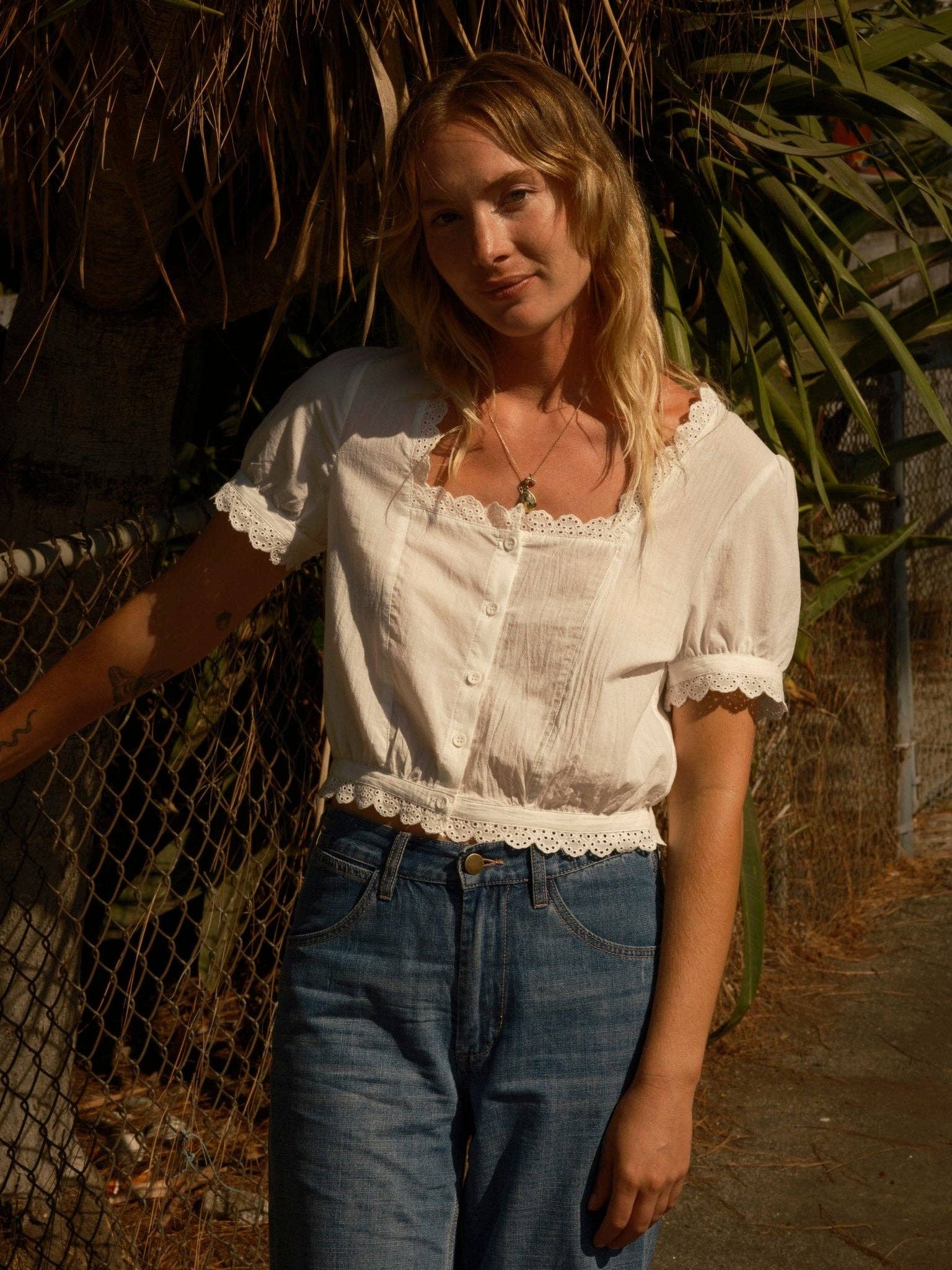 Woman wears a white square-neck top with scalloped eyelet trim, standing by a fence in sunlight.