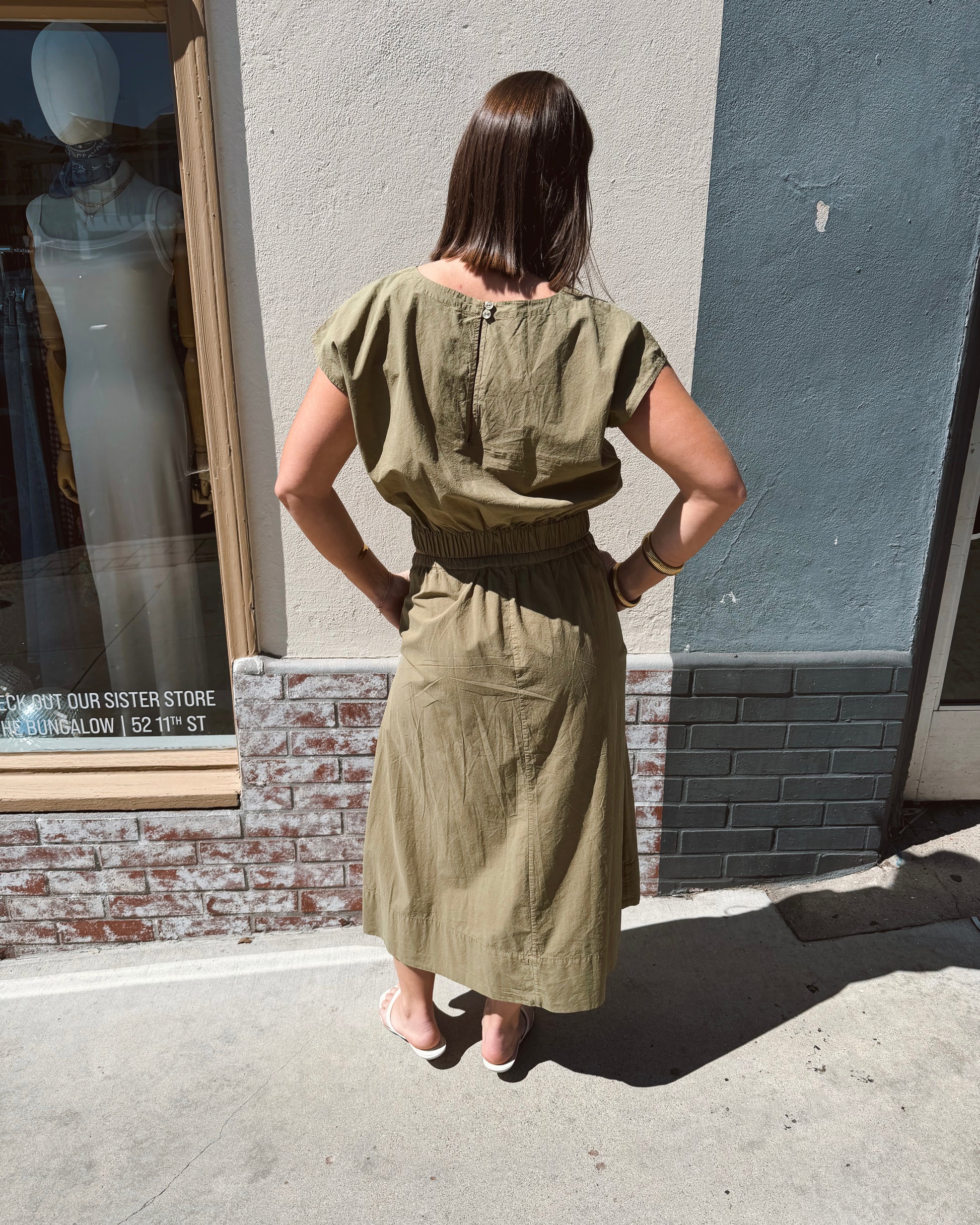 Woman in a dark olive tie-dye Wendy top stands with hands on hips outdoors on a sunny sidewalk.