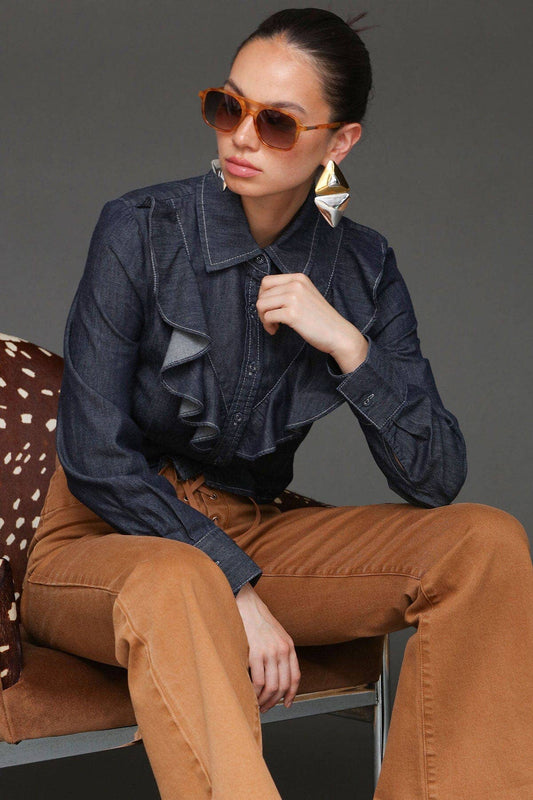 Seated woman in sunglasses, blue denim cropped shirt, statement earrings, and tan pants.