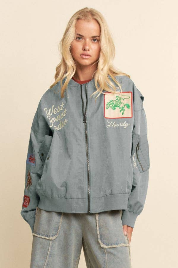 Woman with long blonde hair in an embroidered bomber jacket and light blue jeans.