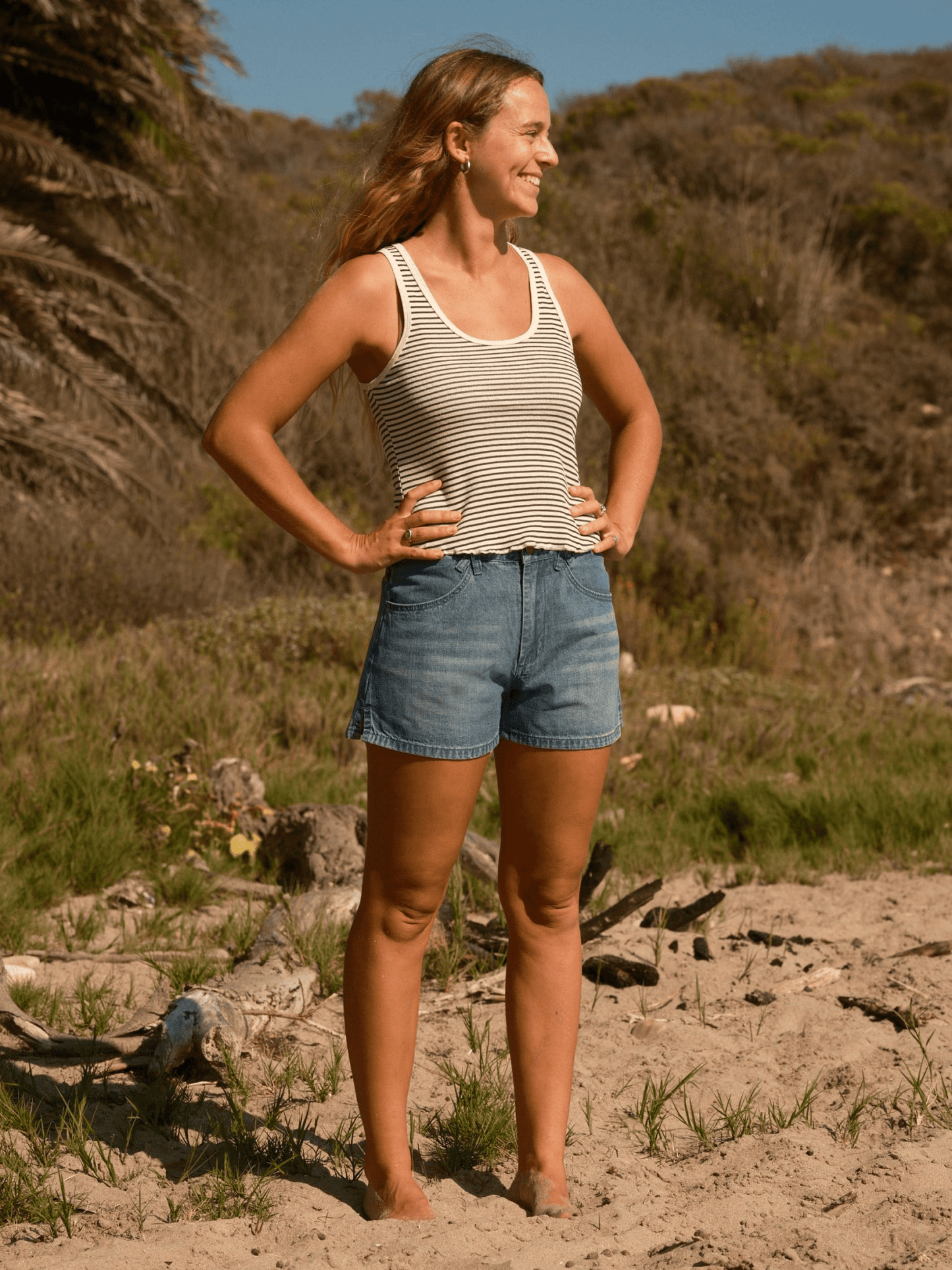 Woman on beach wearing cotton truckie shorts and striped tank top, greenery behind her.