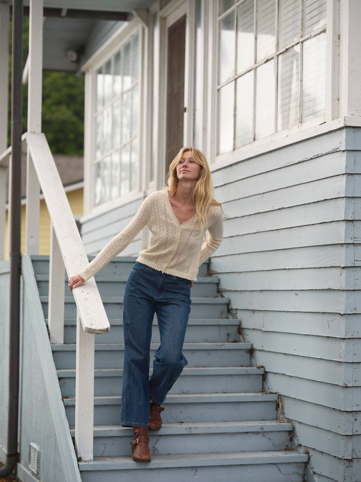 Woman in a beige Juliette cardigan and jeans on blue stairs beside a light blue building. Sweater appears soft and casual.