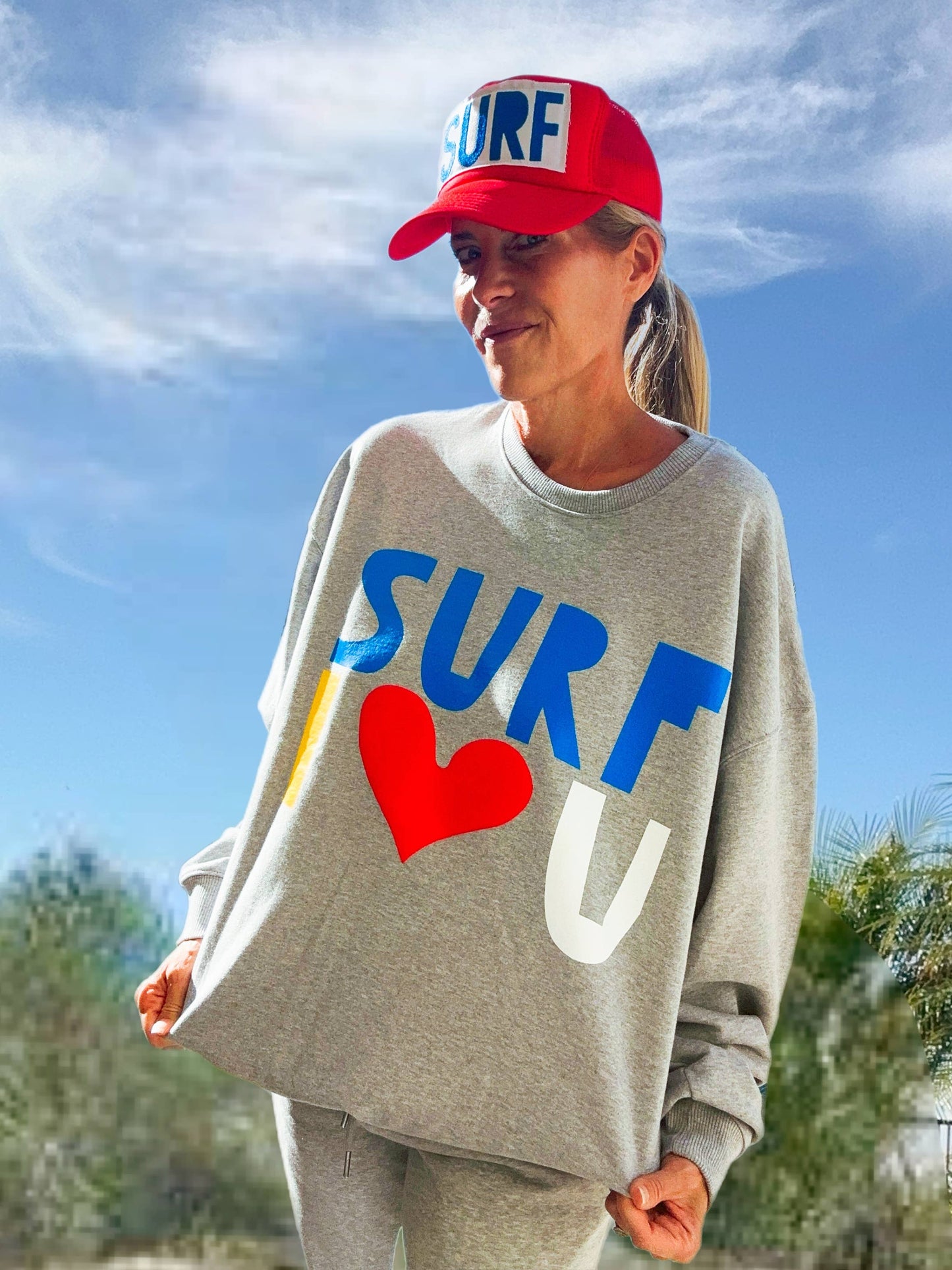 Woman outdoors in a white crewneck sweatshirt and red cap, standing under a blue sky.