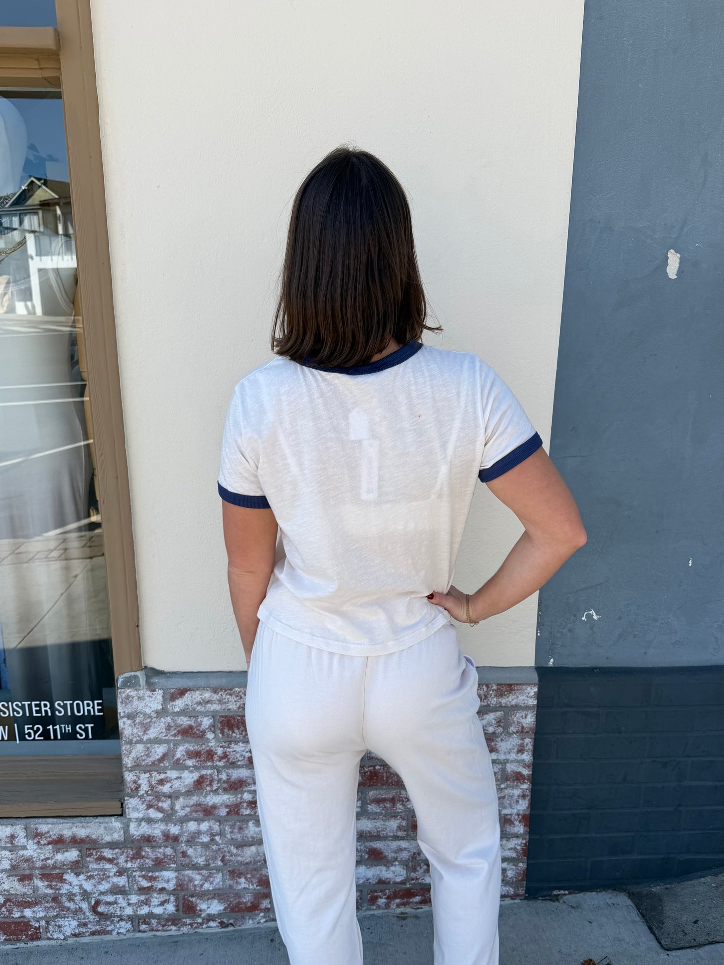 Back view of a person in an oyster-colored boxy ringer crew tee and white pants standing before a brick wall.