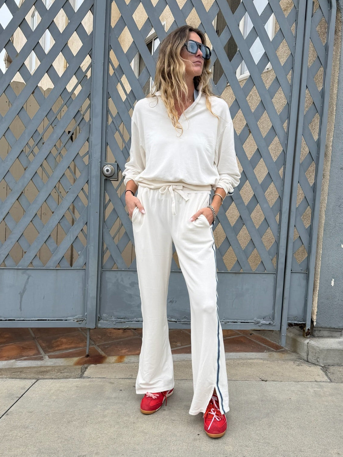 Woman in cream half zip sweatshirt and red sneakers stands with hands in pockets in front of a gray gate.