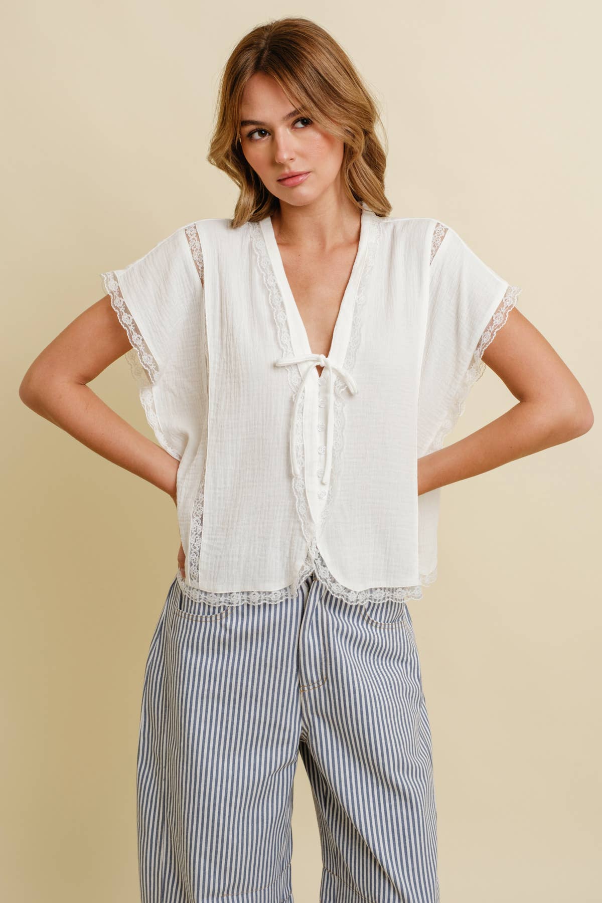 Woman wears a cloud blue cotton top and striped pants, hands on hips, standing before a beige background.