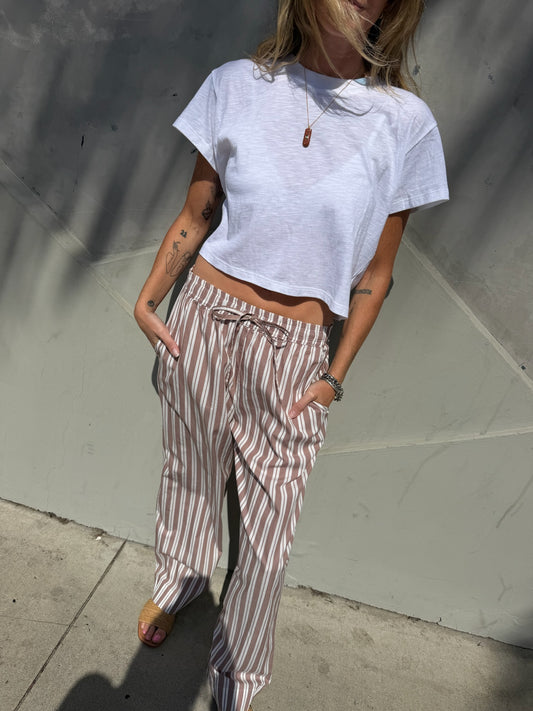 Woman in a cropped white tee and striped wide-leg lounge pants in stucco color stands on a sunny sidewalk, hands in pockets.
