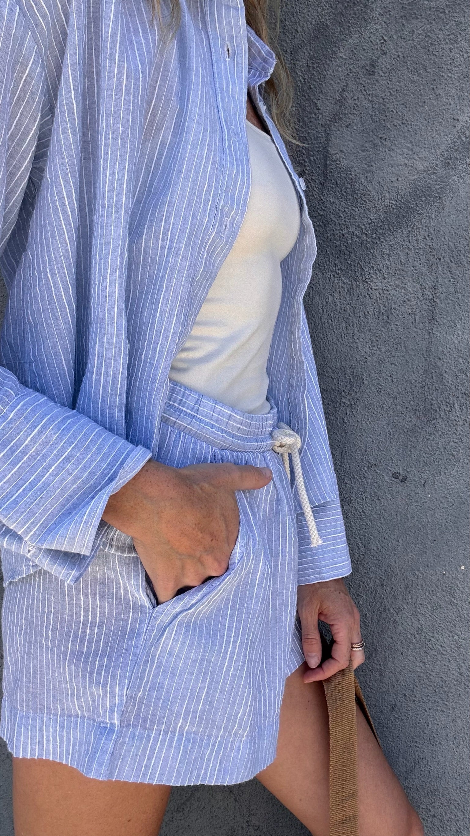 Light blue shorts and a pinstripe shirt over a white tank top, worn by a woman standing beside a gray wall.