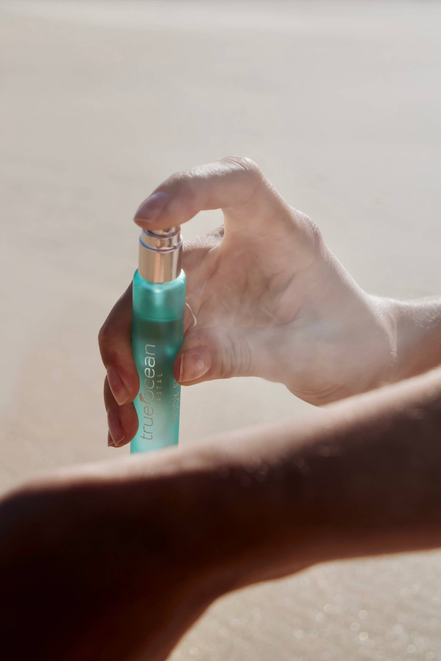 A hand sprays turquoise True Ocean Body Spray onto an arm, emitting a fine mist with a subtle beach scent.