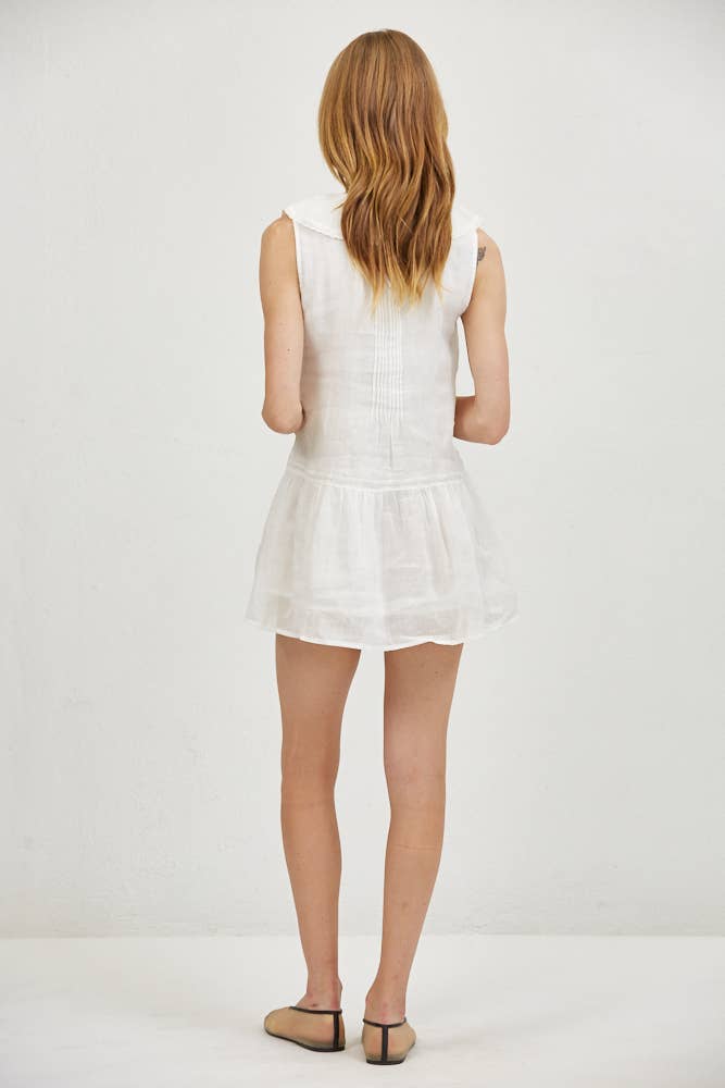 Woman seen from behind in a sleeveless white linen mini dress and flats, standing against a plain wall.
